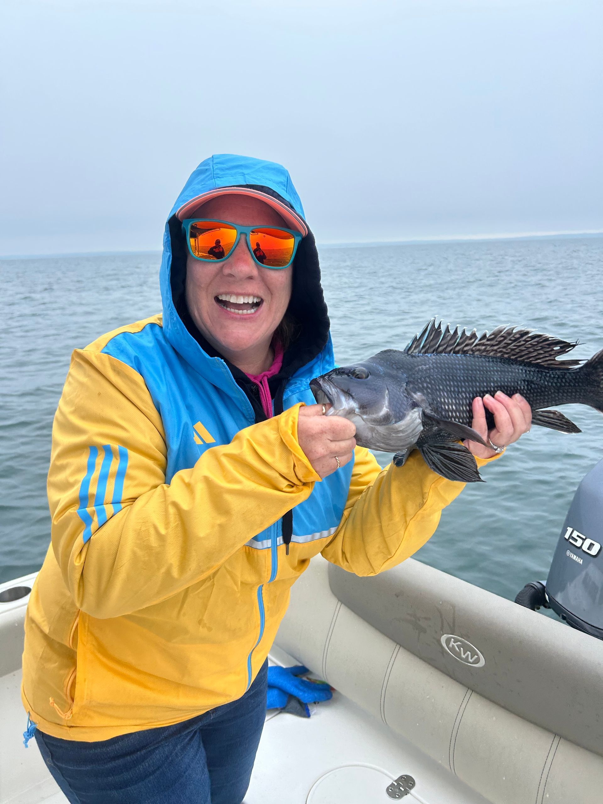 Woman in yellow jacket holds a dark fish on a boat, smiling, with a foggy ocean backdrop.