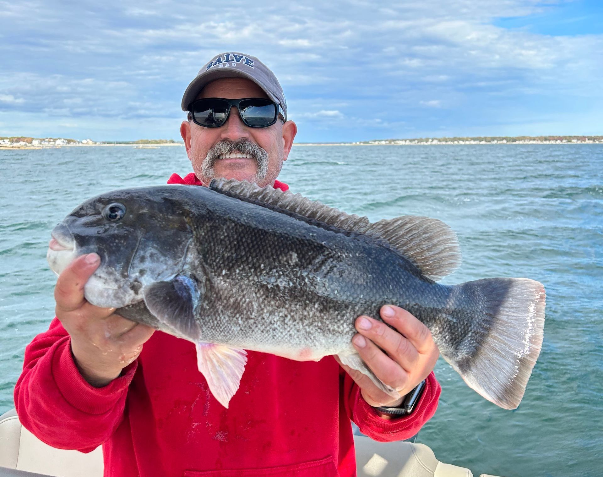 Man holding a large, dark fish he caught; cloudy sky and water in the background.