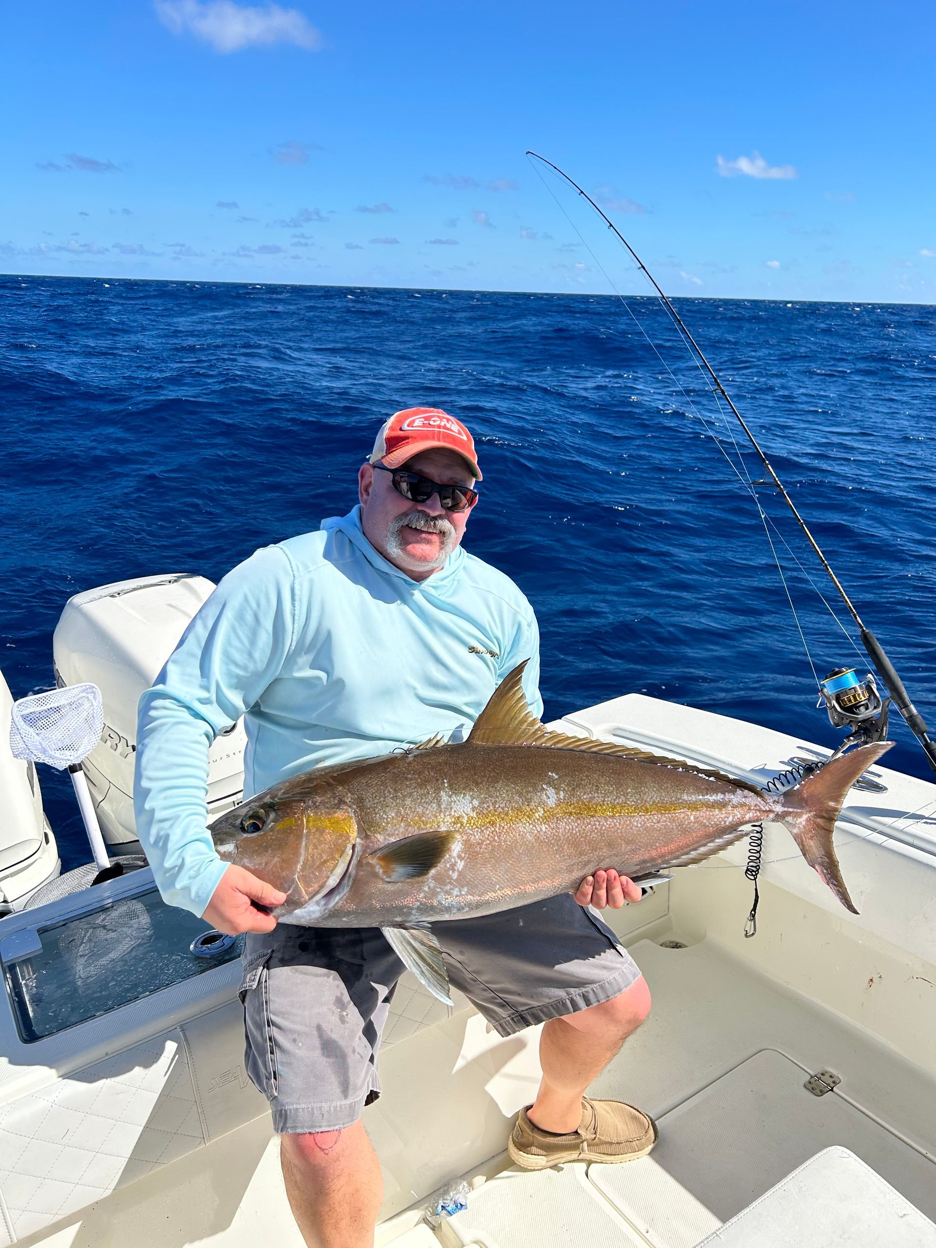 Man on a boat holds a large amberjack fish. He wears sunglasses, a hat, and a light blue shirt. Blue ocean.