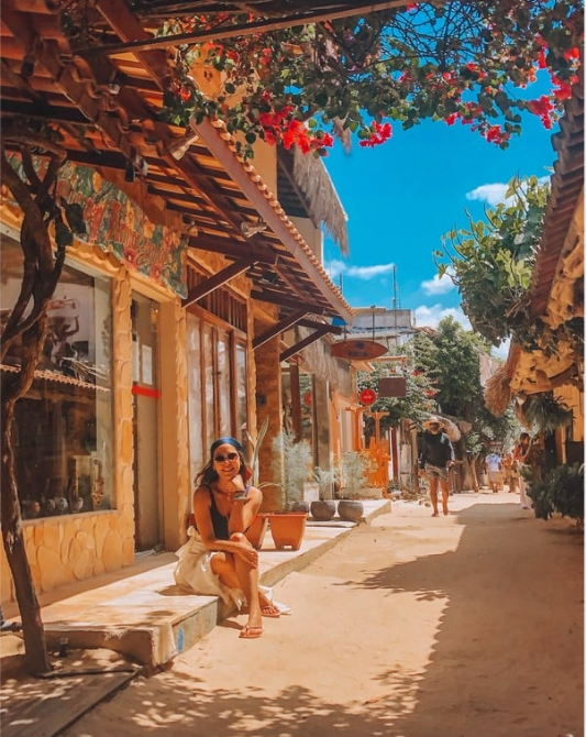 Mulher sentada em uma mureta sorri, viela com lojas e vegetação acima, céu azul.