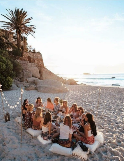 Grupo de pessoas sentadas na praia, desfrutando de uma refeição em volta de uma mesa baixa;
