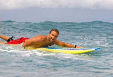 Homem em uma prancha de surfe amarela remando no oceano, vestindo shorts vermelhos e sorrindo.