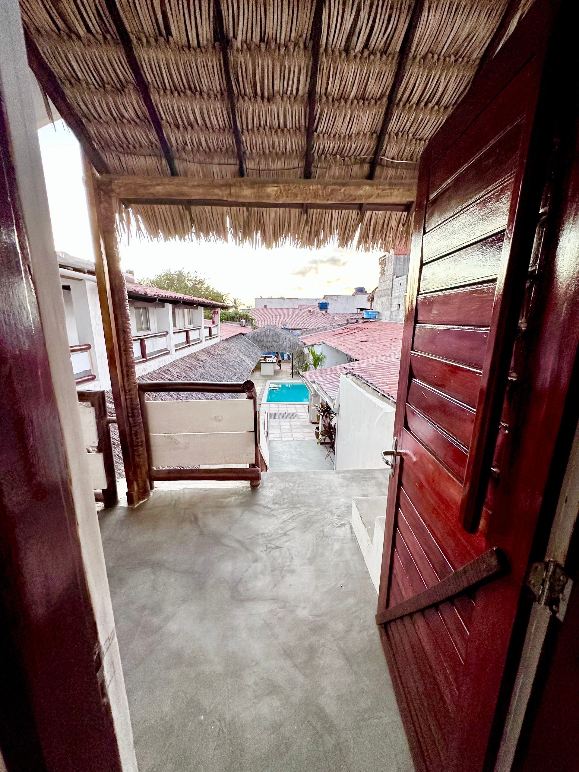 Vista de uma porta para uma varanda com piscina, prédios e telhado de palha.