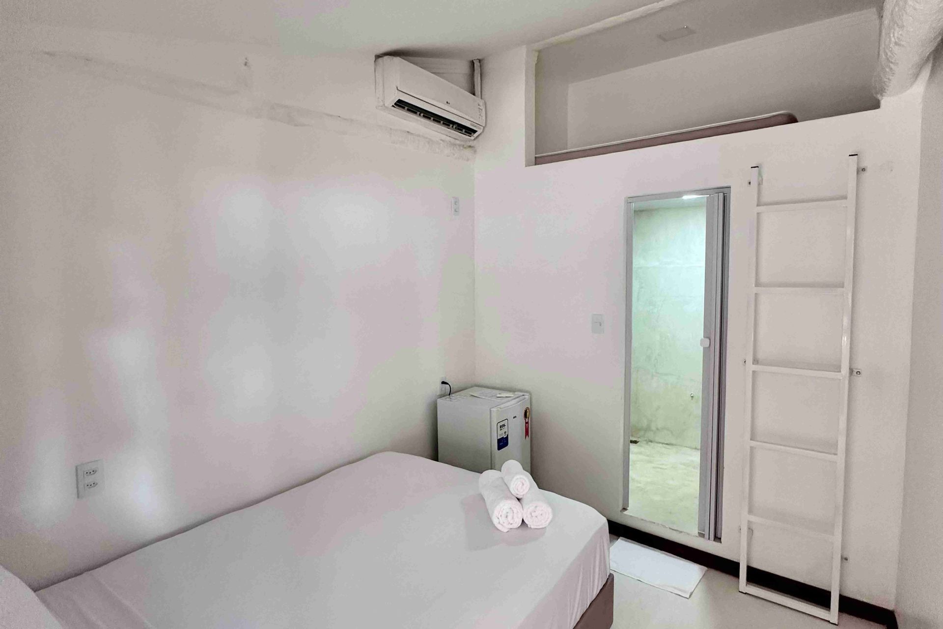 Quarto pequeno, com paredes brancas, cama, frigobar e escada para um mezanino. Uma porta aberta dá acesso ao banheiro.