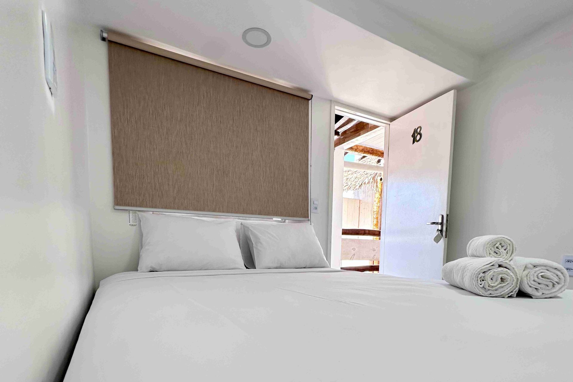 Quarto branco com cama, toalhas enroladas, porta aberta para o exterior e janela com persiana.