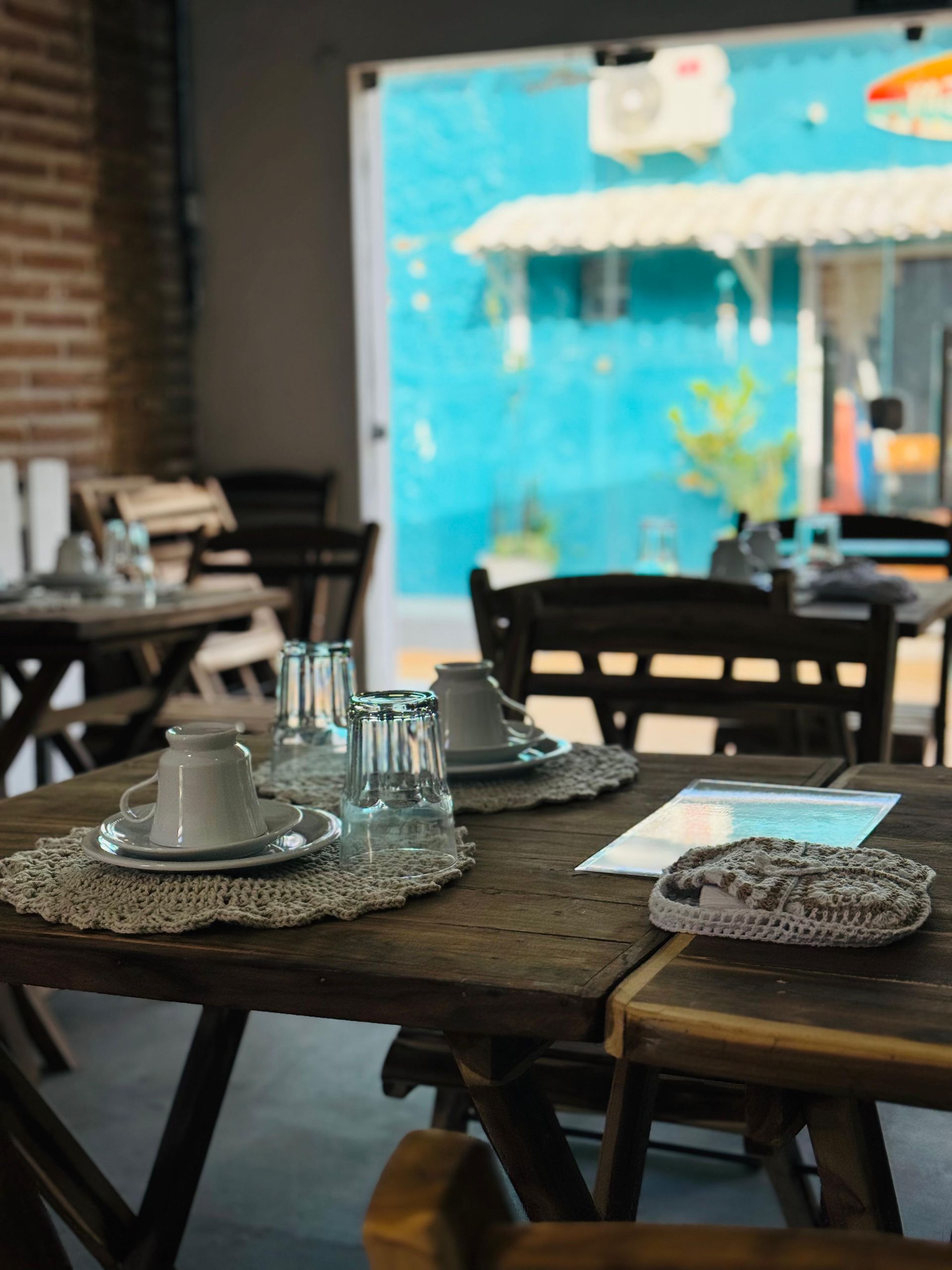 Interior de um café com mesas de madeira decoradas com xícaras, copos e toalhinhas de crochê.