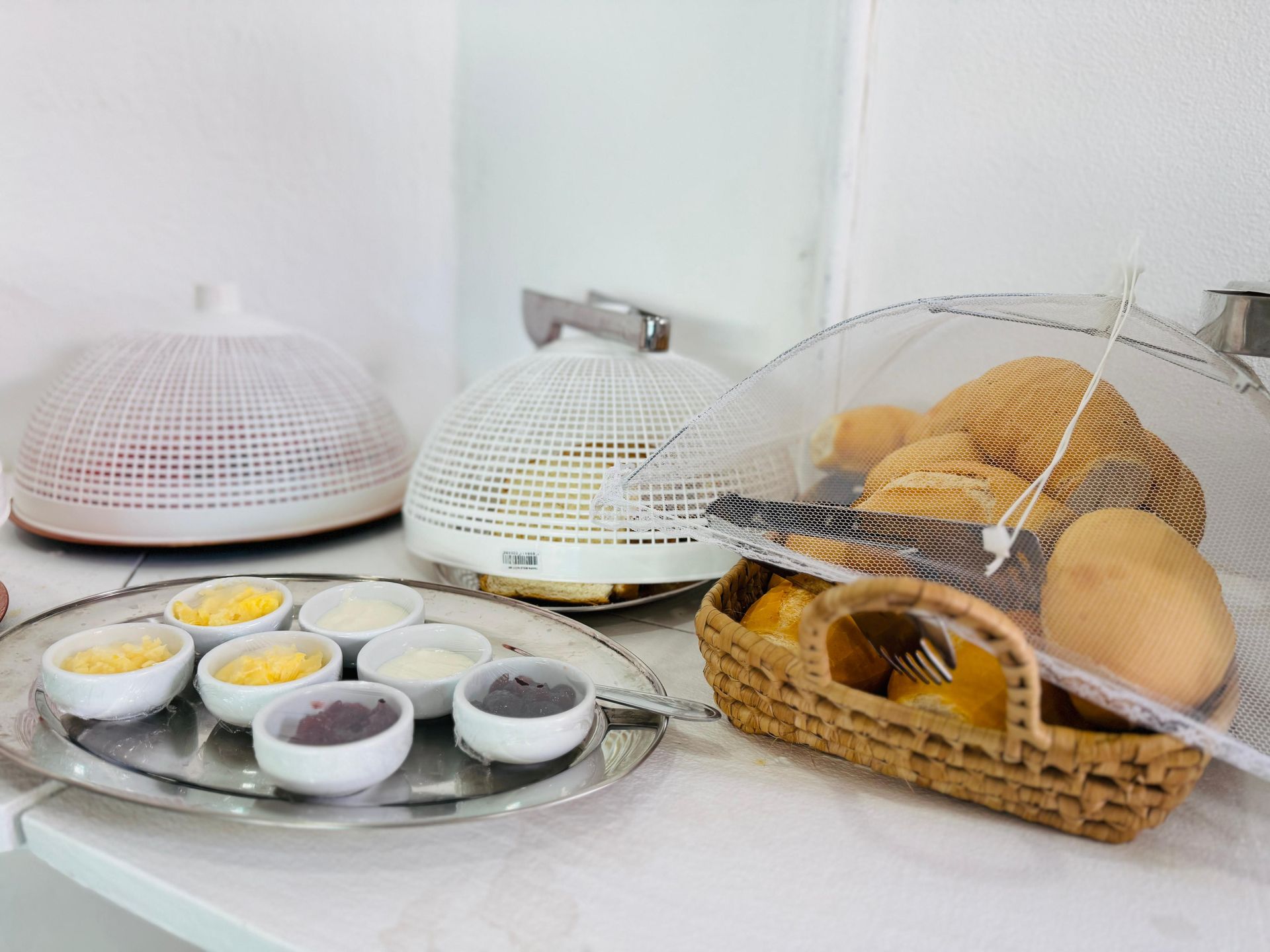 Buffet de café da manhã com pães, acompanhamentos e frutas, coberto por uma tela.