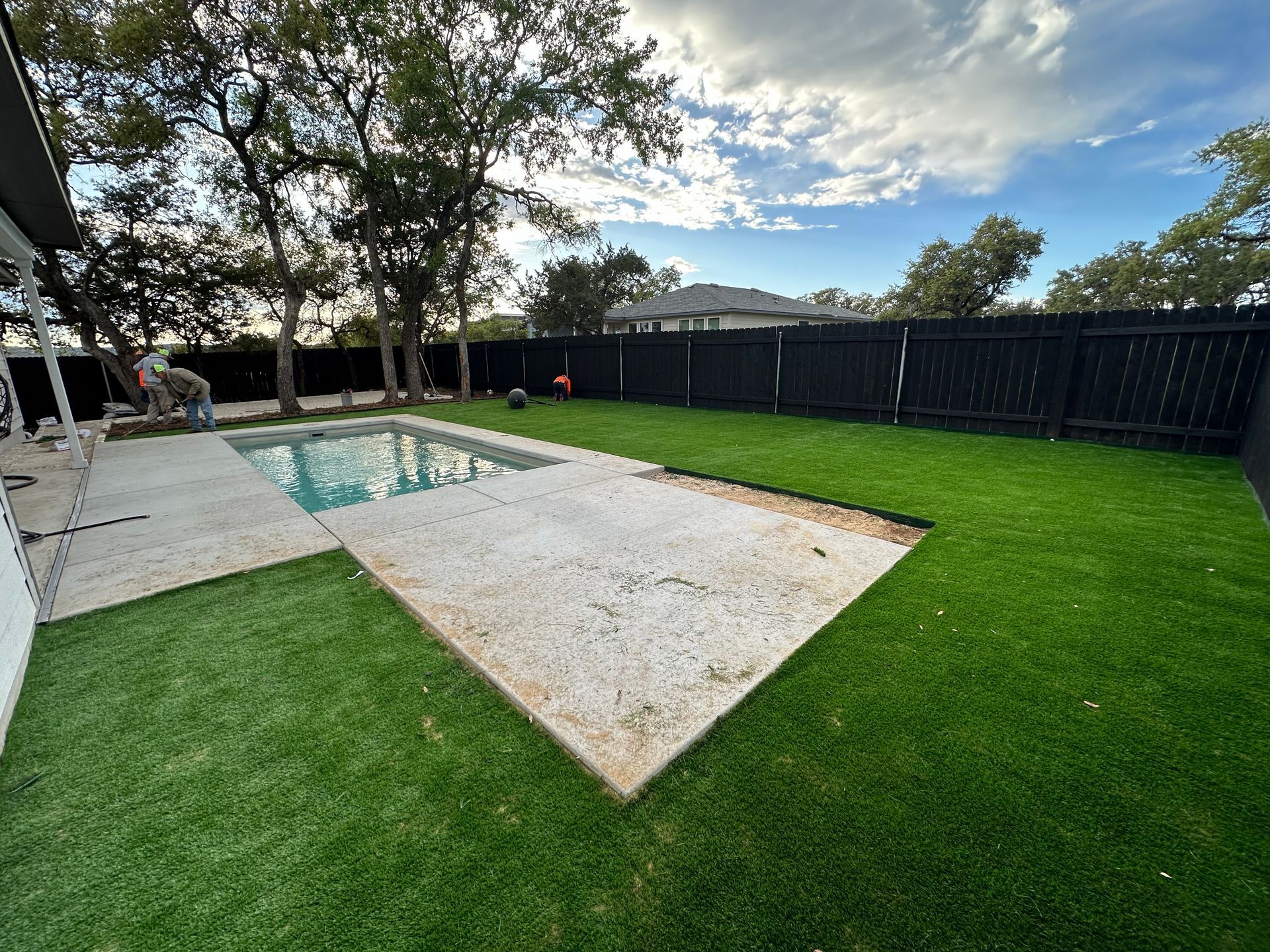 Backyard pool area with white patio, green lawn, black fence, and trees under a partly cloudy sky