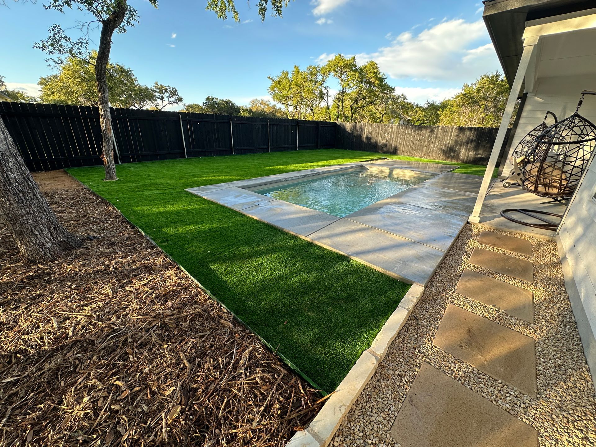 Backyard with a swimming pool, green lawn, and dark fence under a blue sky.