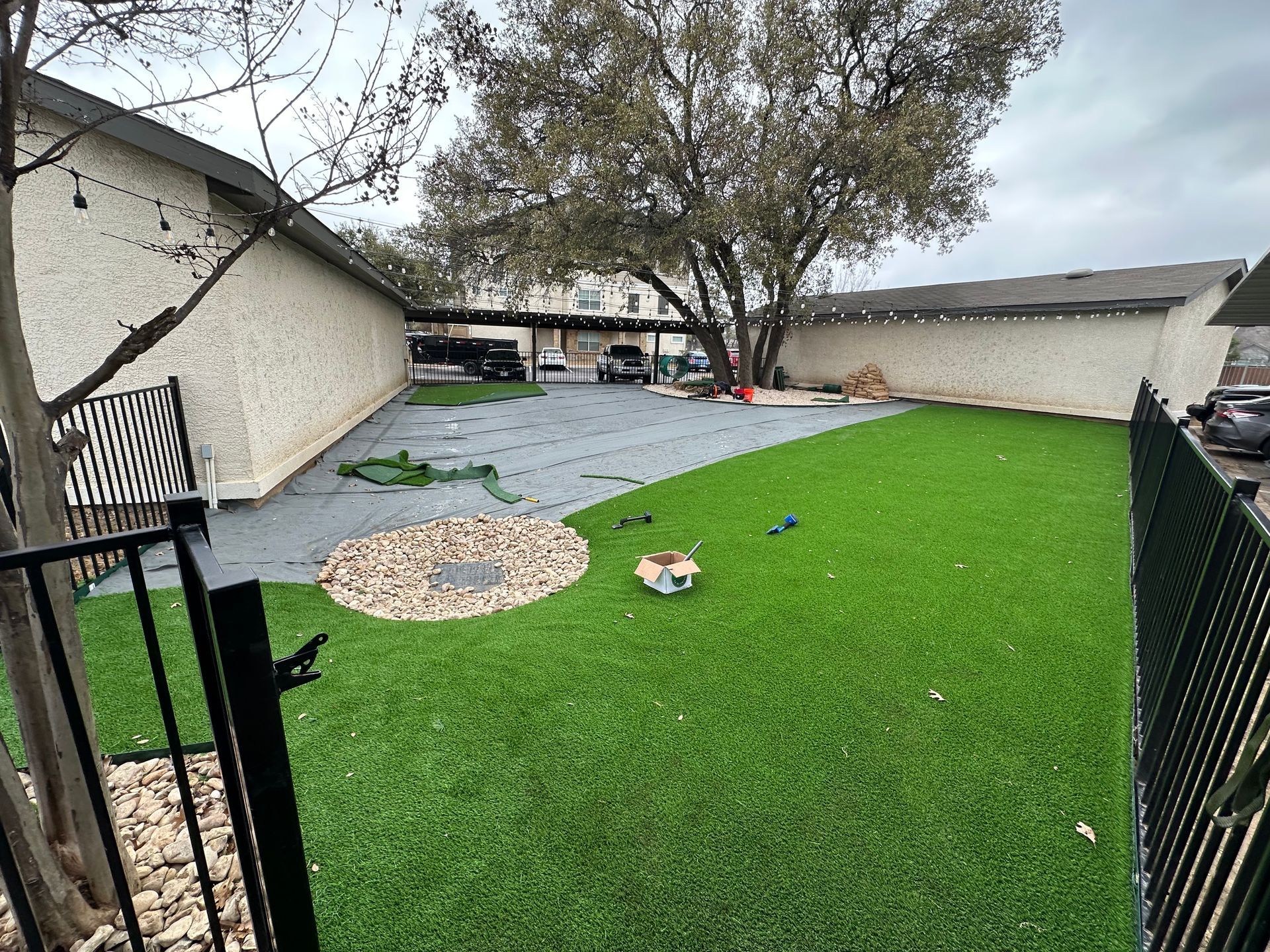 A backyard space featuring a green artificial grass lawn, a grey patio area, a rock garden circle, and black metal fencing.