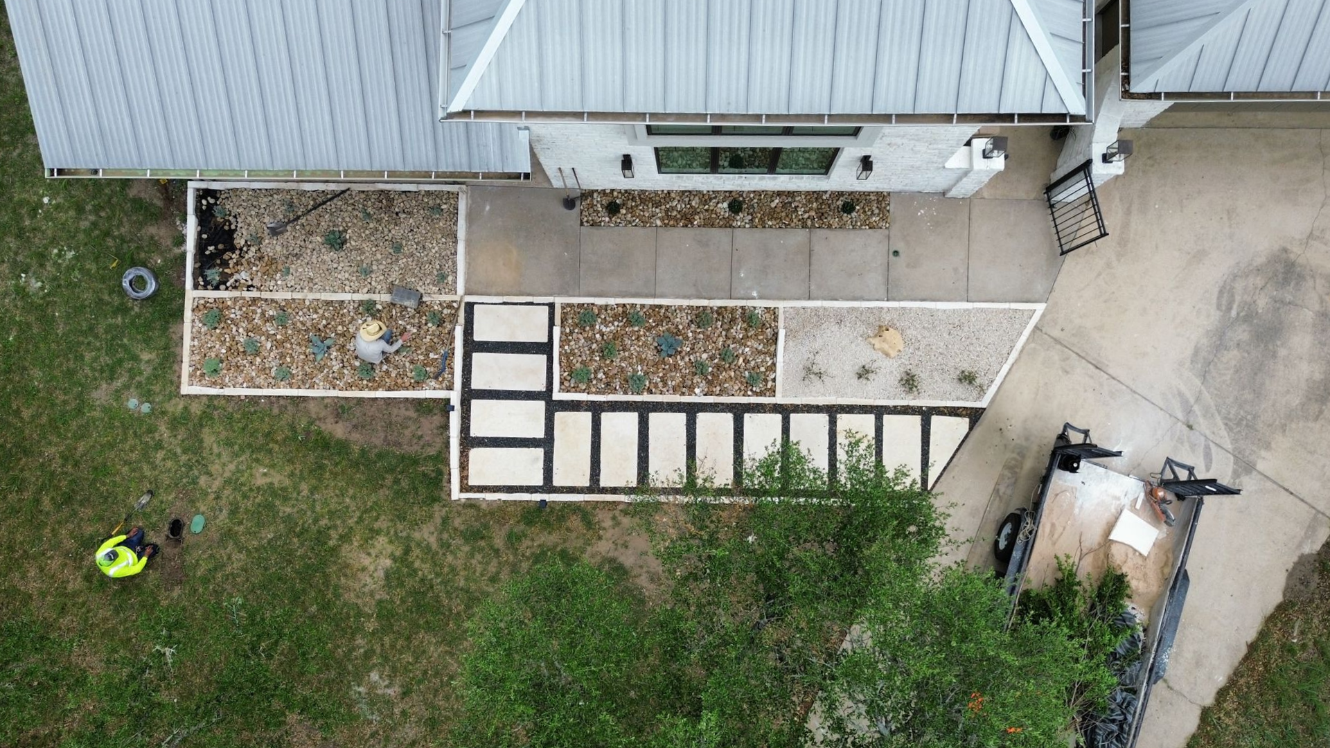 Overhead view of a house's entrance with landscaping, concrete pathway, gravel, and a person in a yellow vest on the lawn.