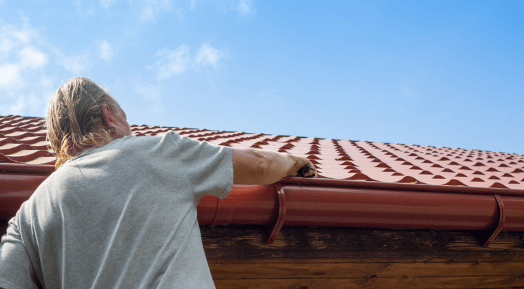 A man is standing on a roof looking at a gutter.