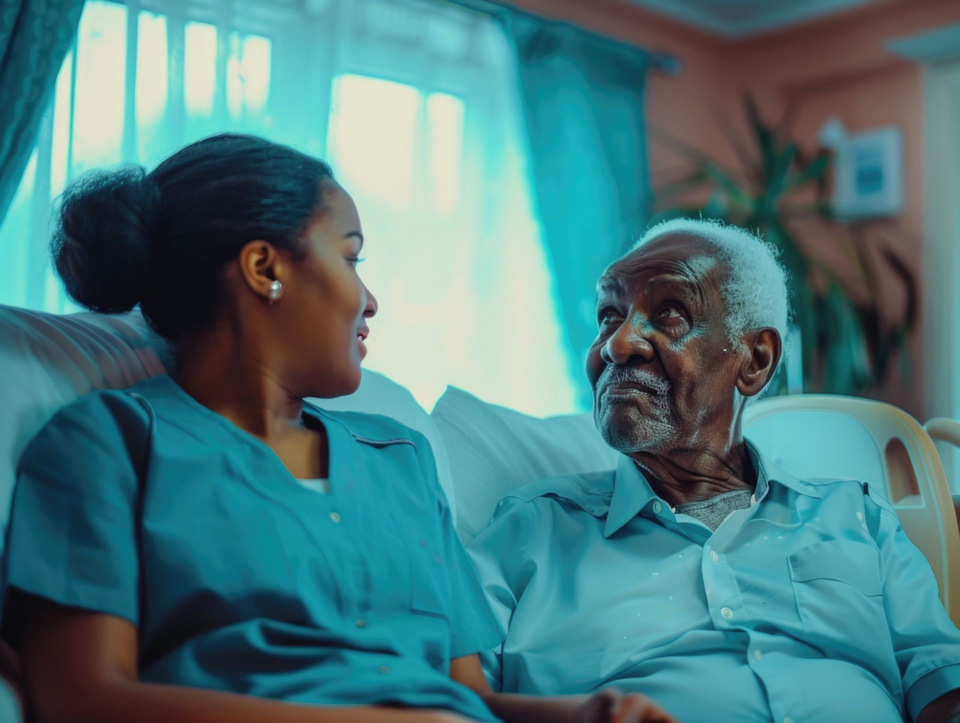Woman in blue scrubs smiles at elderly man; they sit indoors.