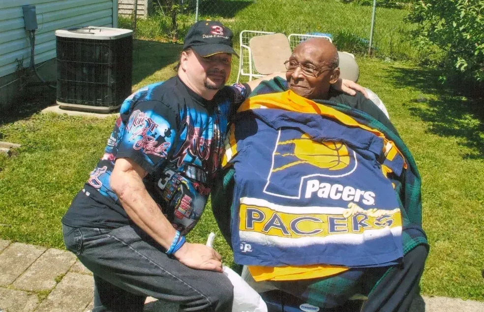 Man in cap and shirt with elderly person wrapped in a Pacers jersey outside.