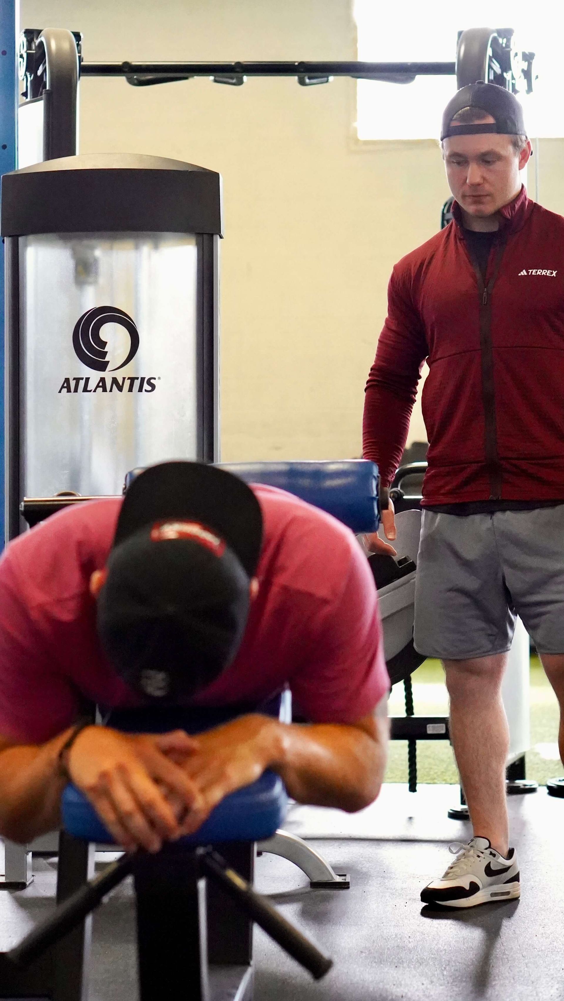 A man is sitting on a bench in a gym while another man stands behind him.