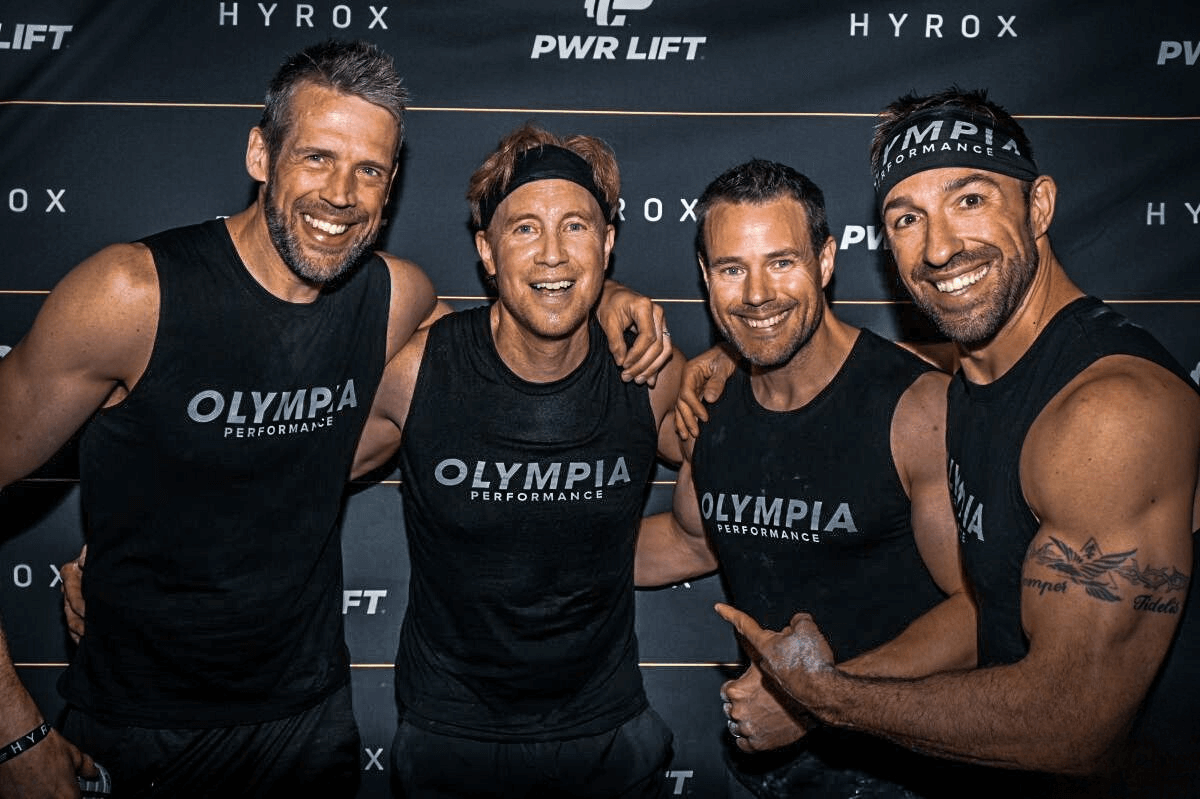 Four men are posing for a picture in front of a wall that says Olympia Performance