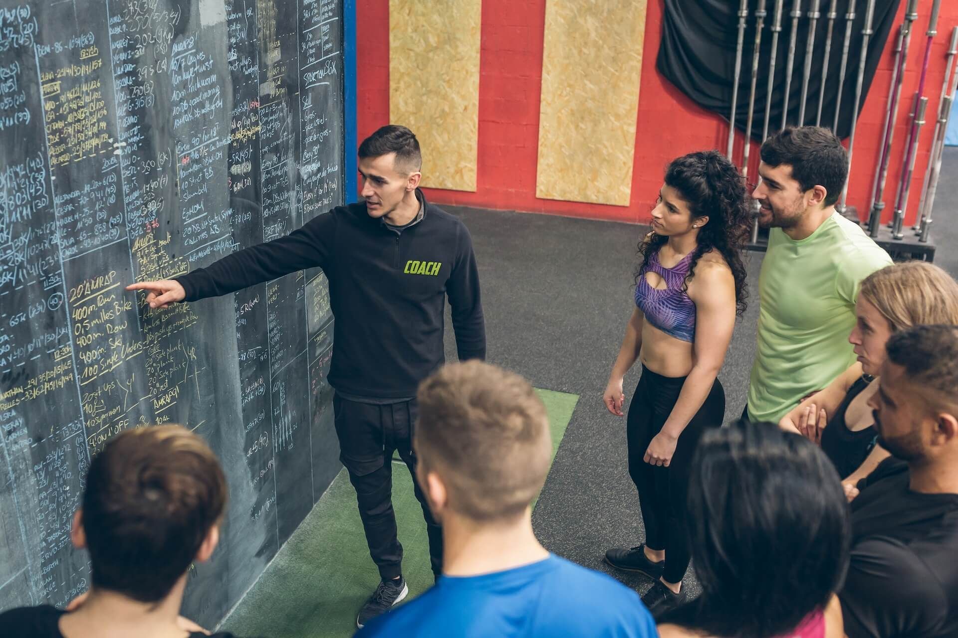 Instructor points to a chalkboard with workout details as a group of people look on in a gym.