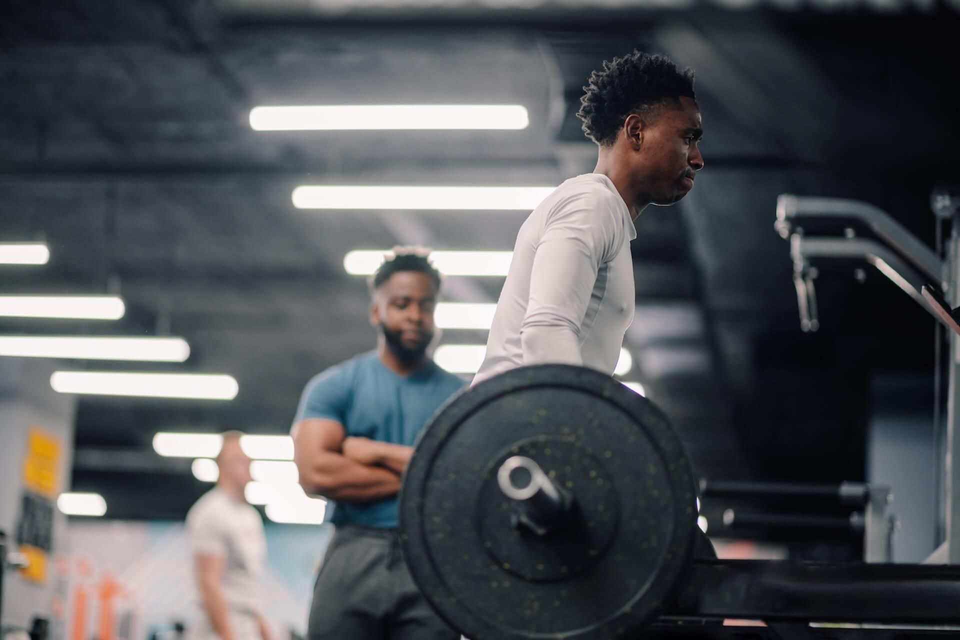 Man lifting barbell in gym, coach watches.