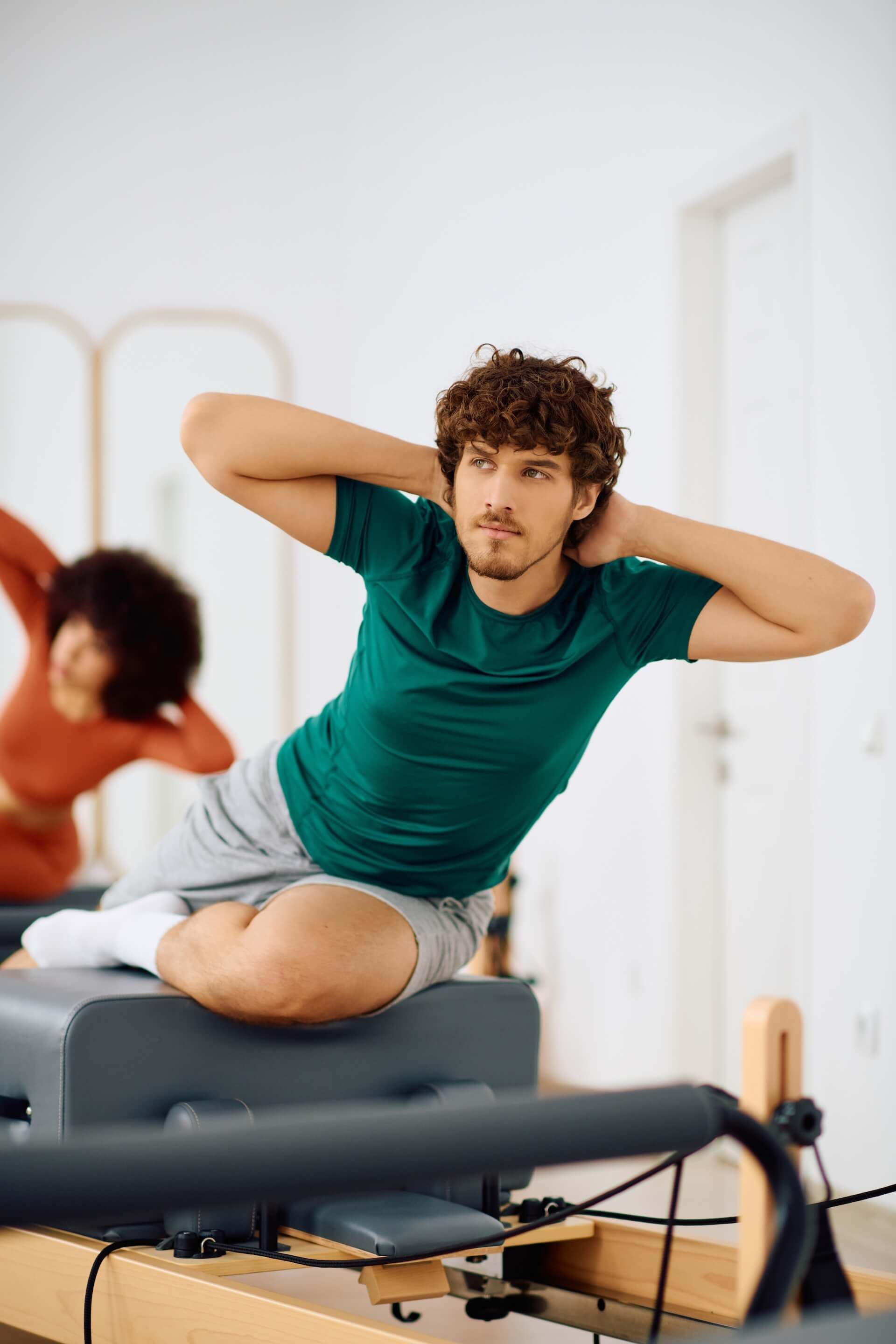Man in green shirt doing Pilates on a reformer, hands behind head; woman in orange shirt in the background.