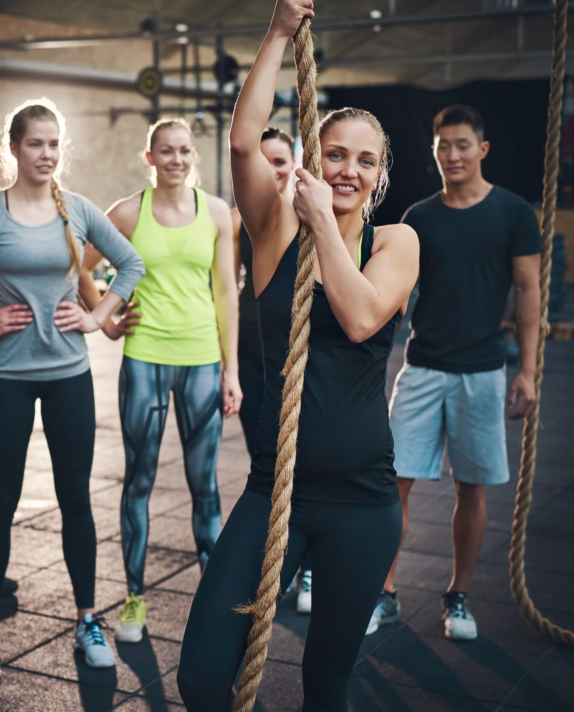 Woman climbing a rope at a gym, smiling, with a group of people watching.