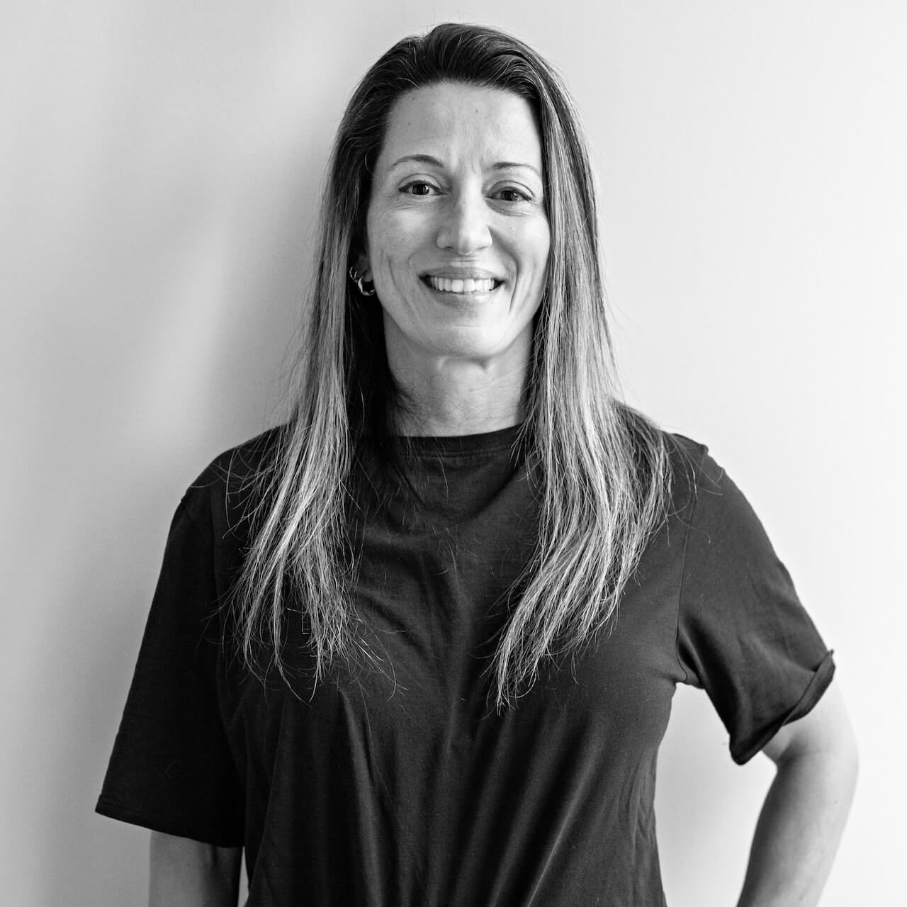 Woman with dark hair, smiling at the camera. Wearing a black and white top, near a white wall.