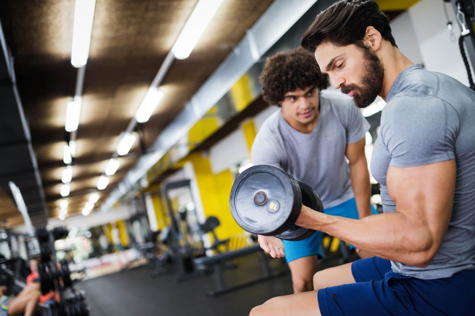 Man lifts dumbbell with trainer's assistance at a gym.