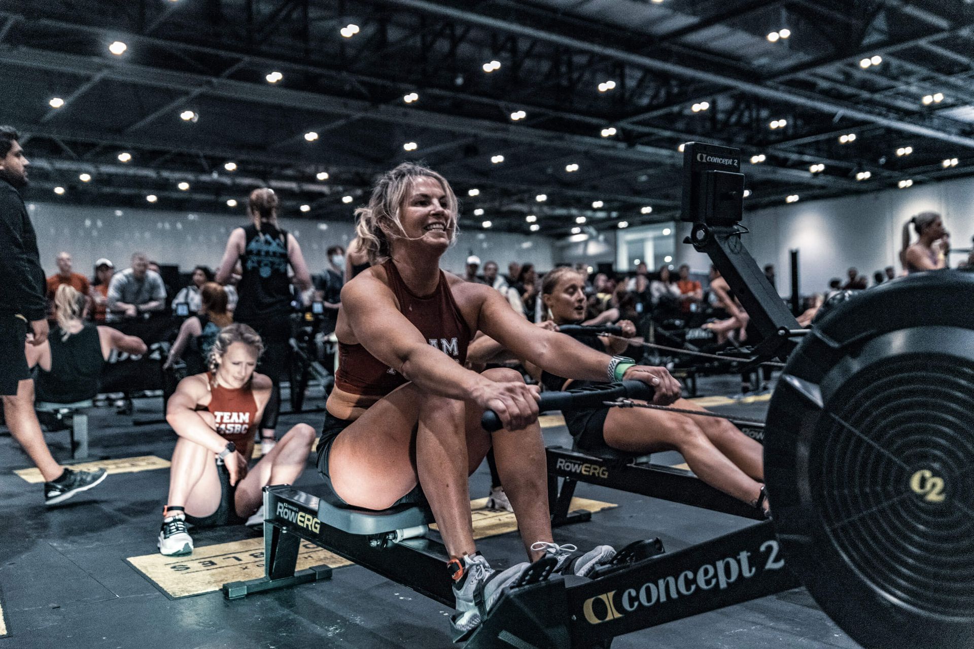 Woman smiling on a rowing machine during a fitness competition; other competitors and audience in the background.