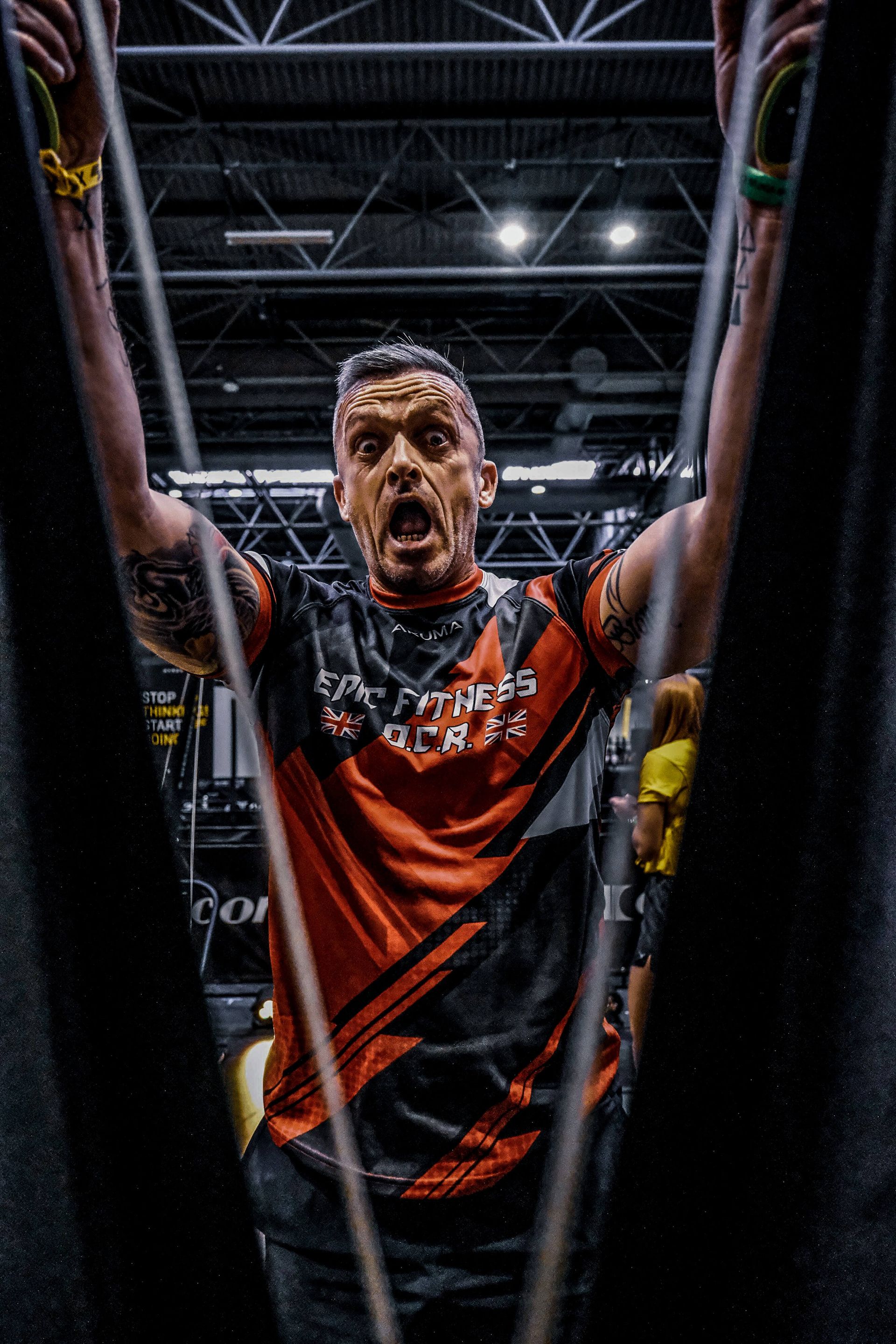 Man in red and black shirt with arms raised, mouth open, framed by dark panels.