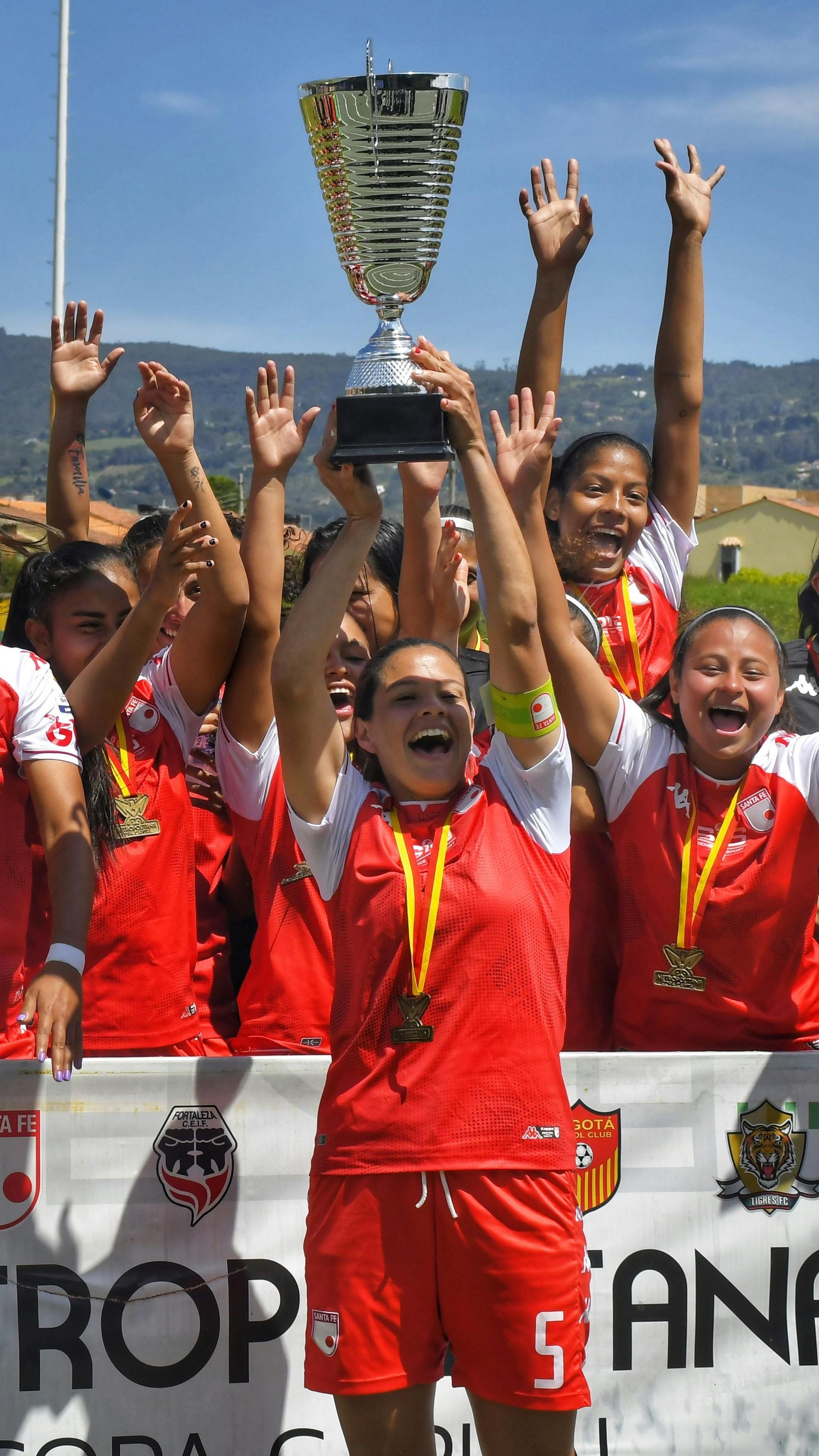 Soccer team in red jerseys celebrating a win, holding up a trophy on a sunny day.