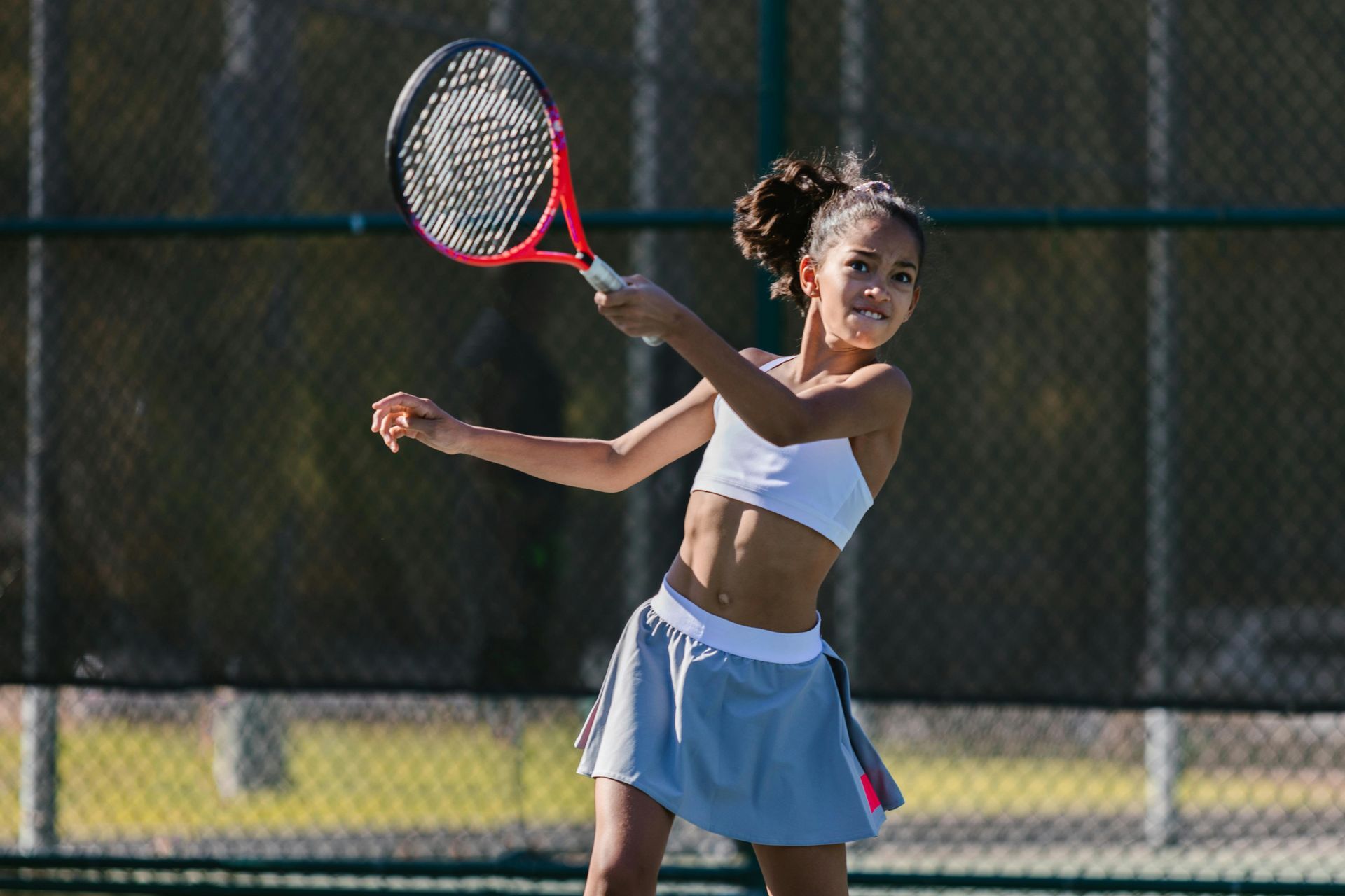 Girl in tennis outfit swings a red racket on a tennis court, ready to hit the ball.