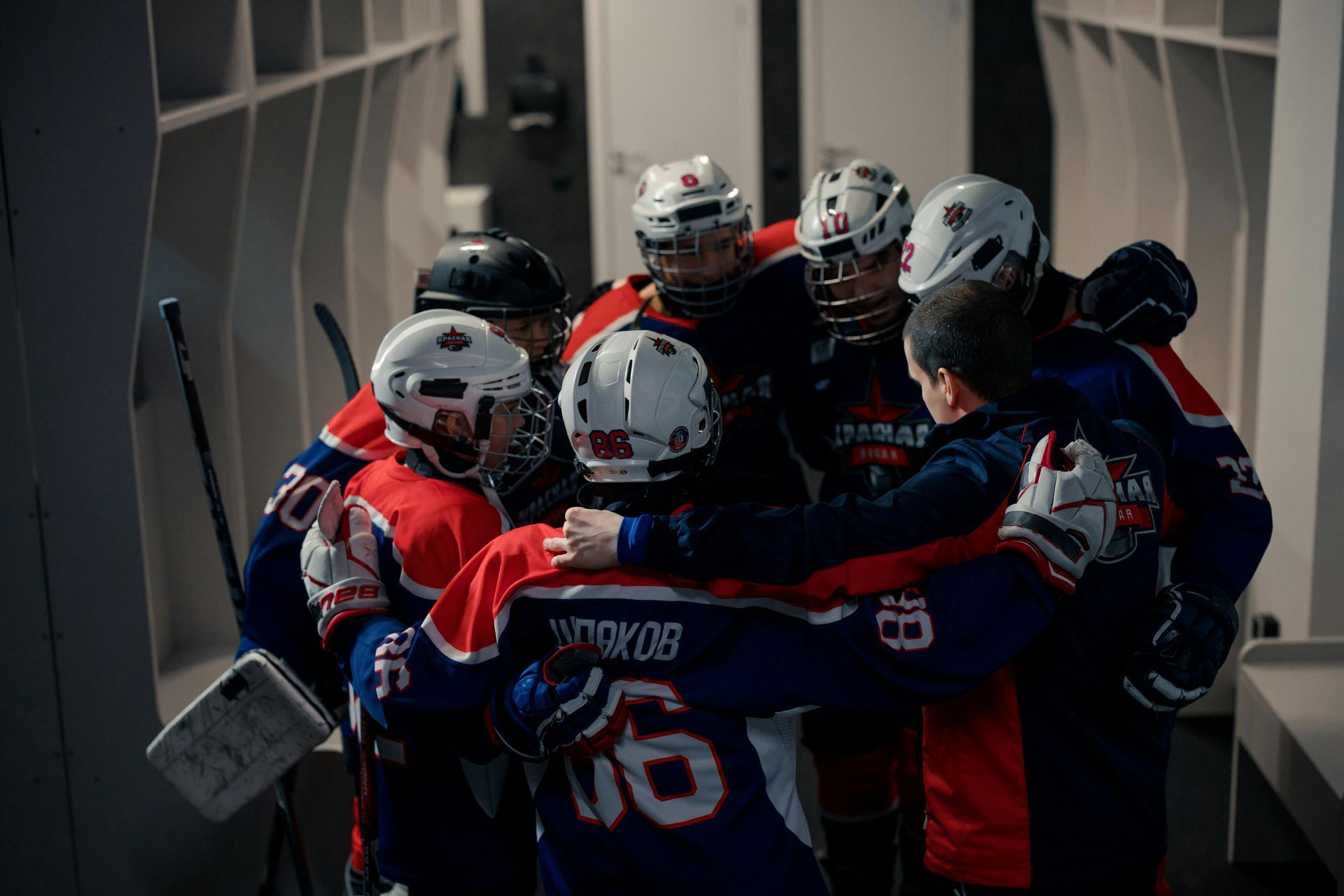 Hockey team huddle in locker room. Red, blue jerseys, white helmets. Arms around each other, focused, supportive.
