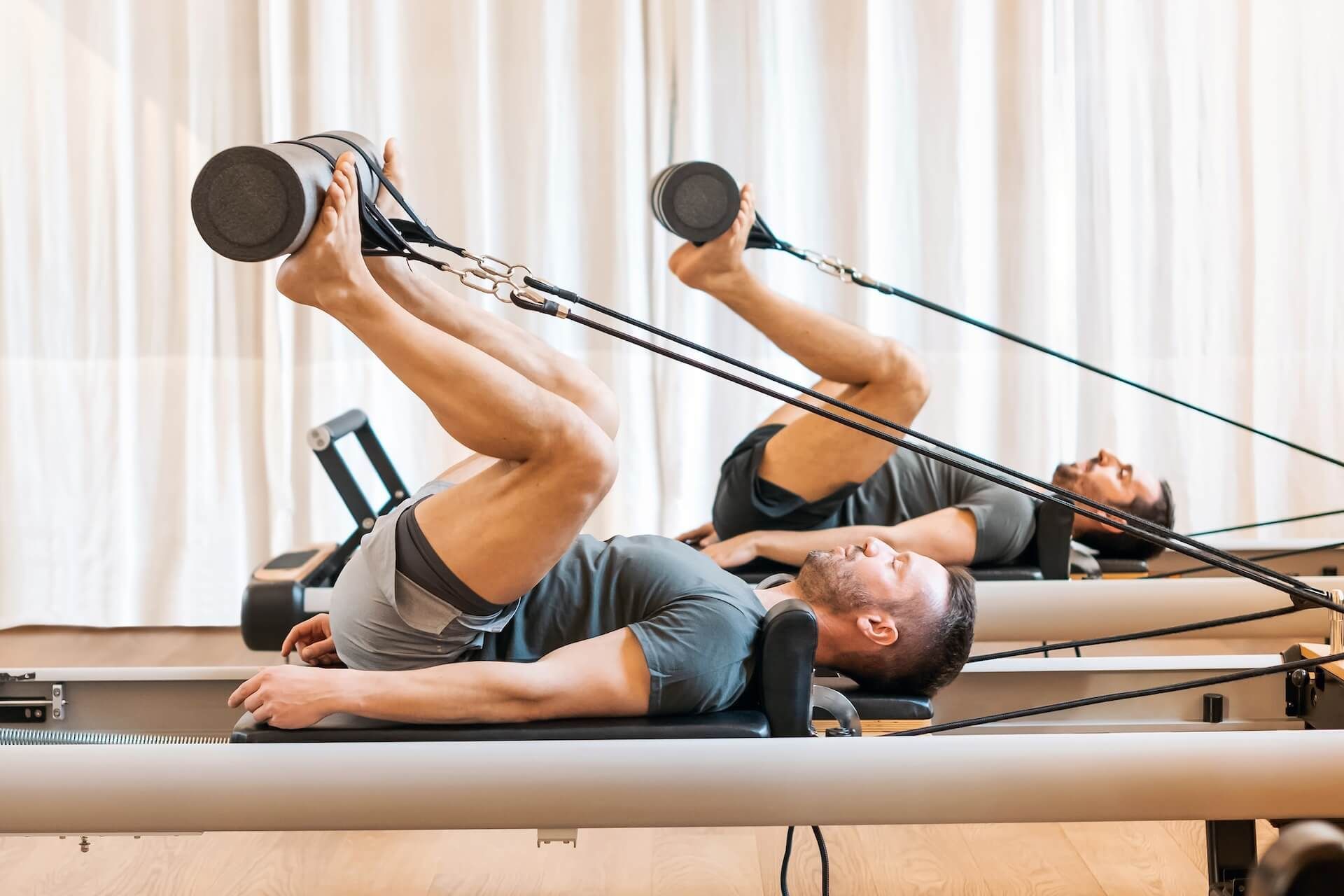 Two men doing Pilates exercises on reformer machines in a studio.