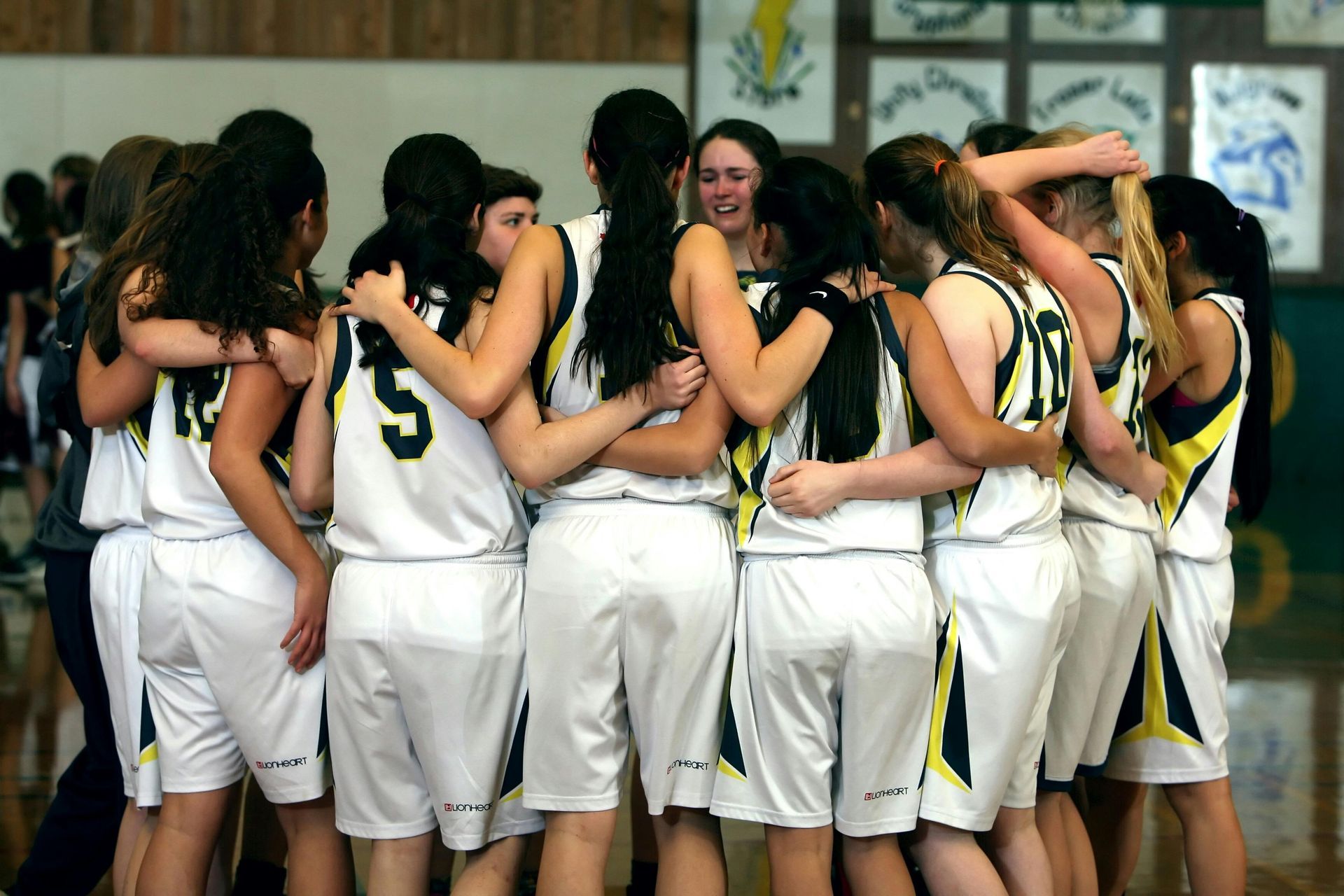 Basketball team huddles together, arms around each other, wearing white and yellow uniforms.