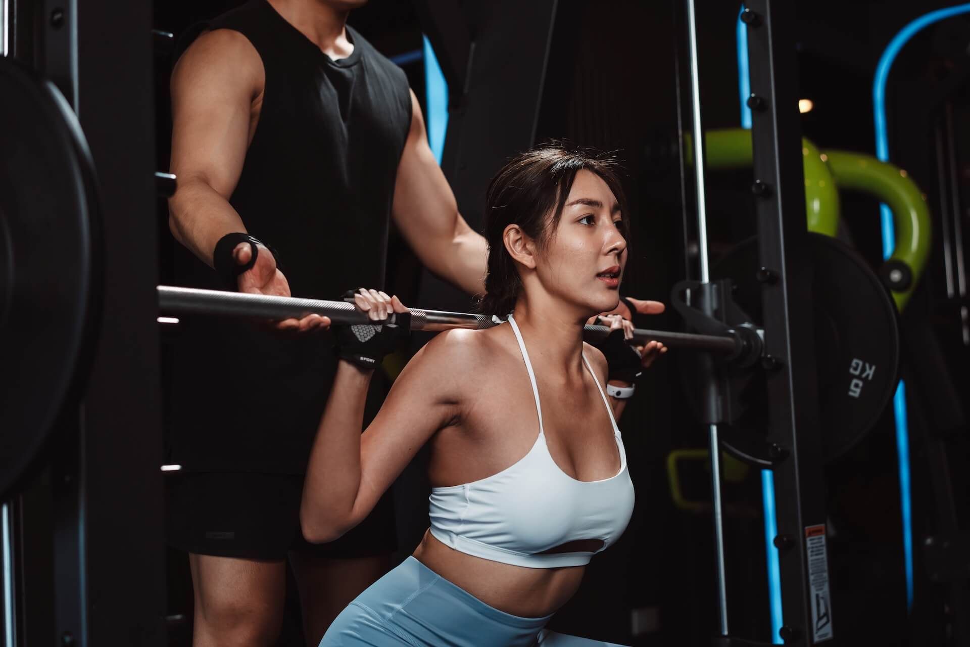 Woman performing barbell squats in a gym, assisted by a spotter.
