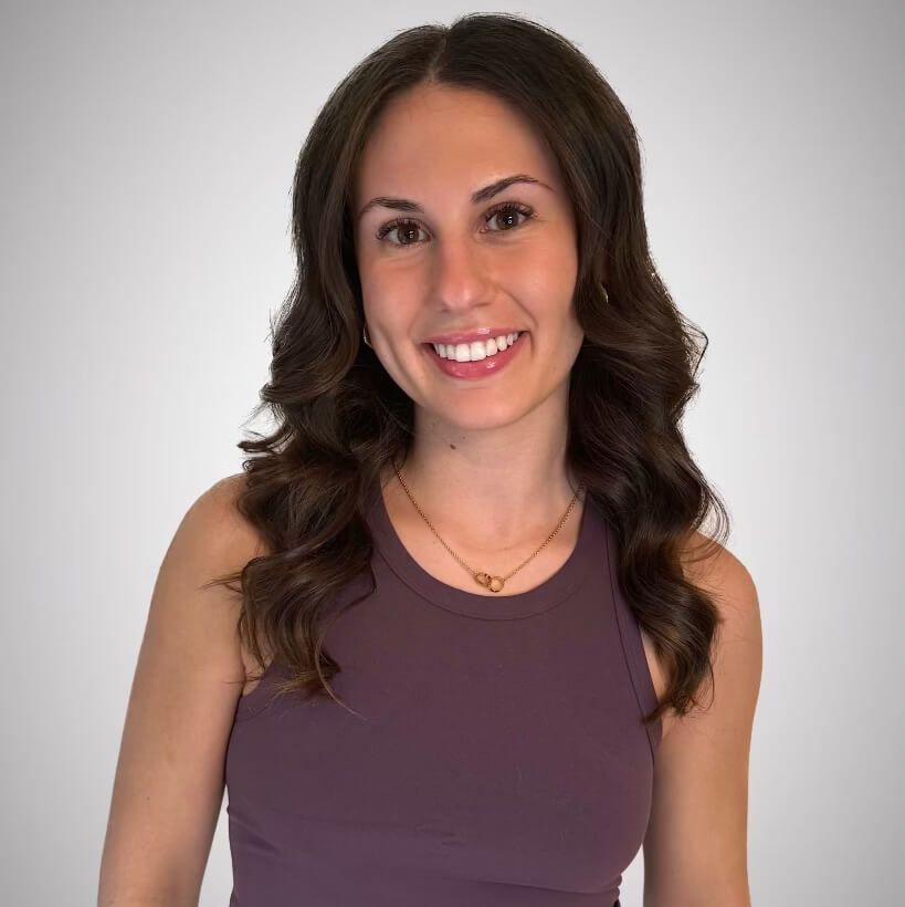 Woman with long, wavy brown hair smiles at the camera, wearing a purple tank top and gold necklace.