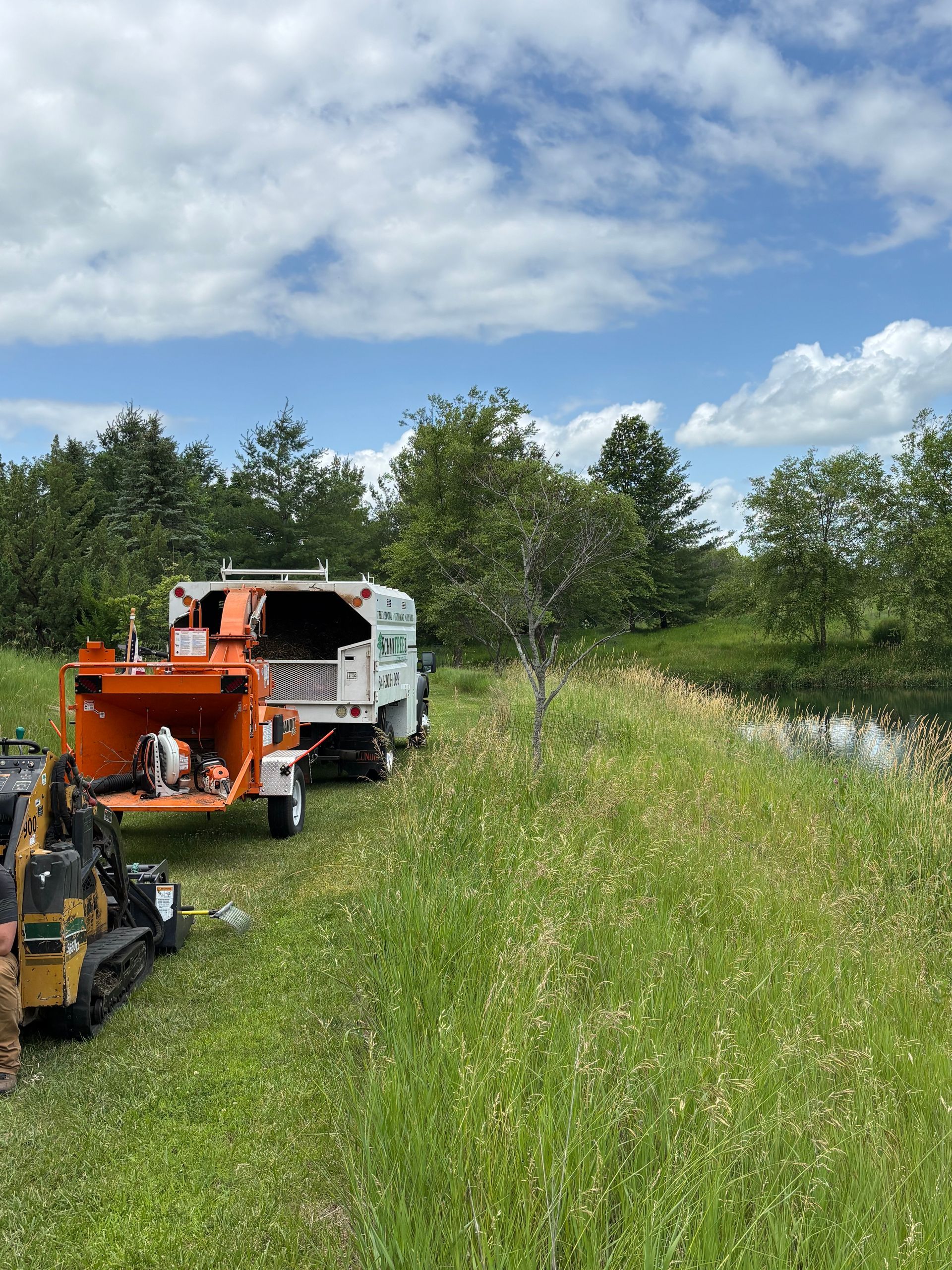 Orange and white wood chipper next to a small excavator in a field with a pond and trees.