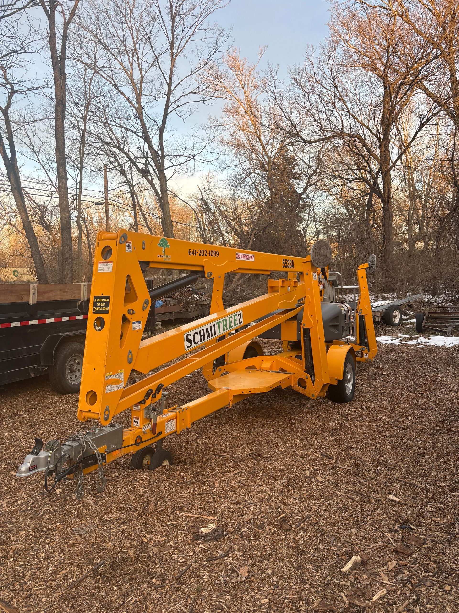 Yellow self-propelled tree lift on trailer, in a yard with wood chips and trees.