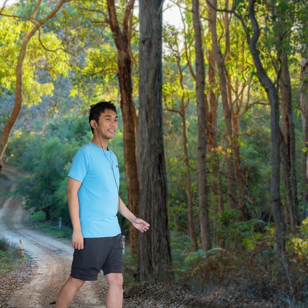 A Man In A Blue Shirt Is Walking Down A Dirt Road In The Woods — Sea & Sand Chiropractic In Toukley, NSW