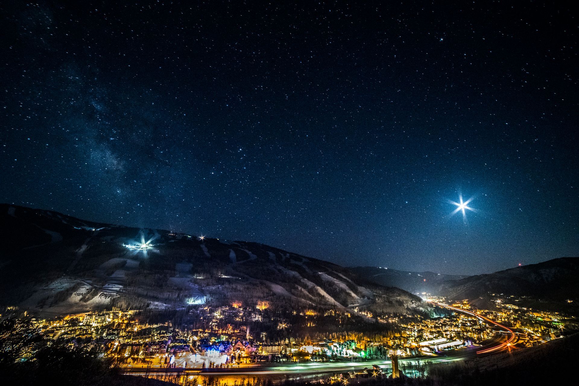 Night sky over a town; stars and bright lights sparkle. Dark hills frame the city with a large, bright star.