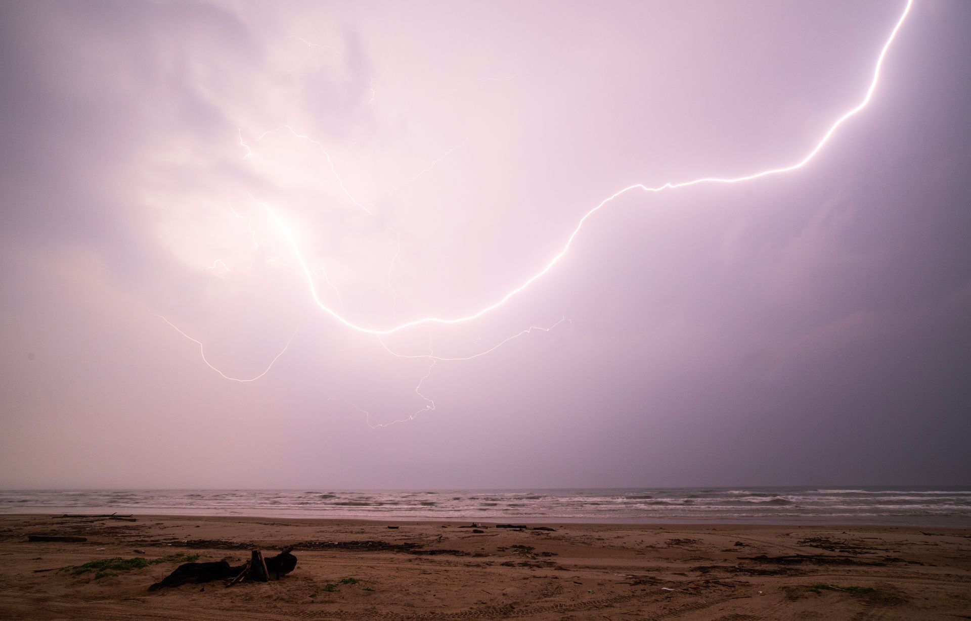 Lightning strikes over a beach and ocean; stormy sky with purplish hue.