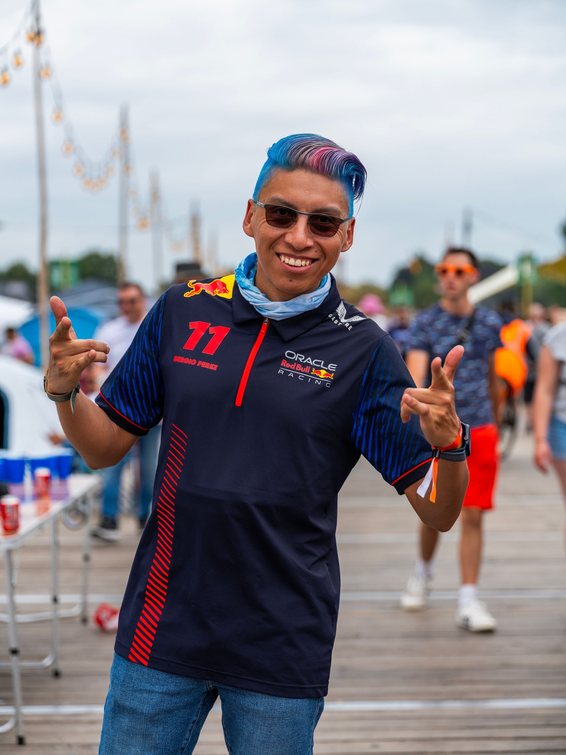 Man with blue hair, Red Bull shirt, pointing and smiling outdoors.