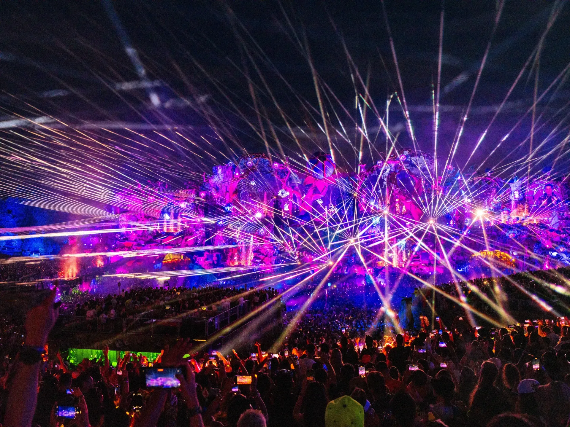 Concert stage with vibrant purple and white lasers, a large crowd, and lights in the night sky.