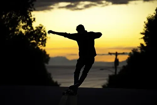 Silhouette of person skateboarding down a road at sunset, arm outstretched for balance, trees on either side.