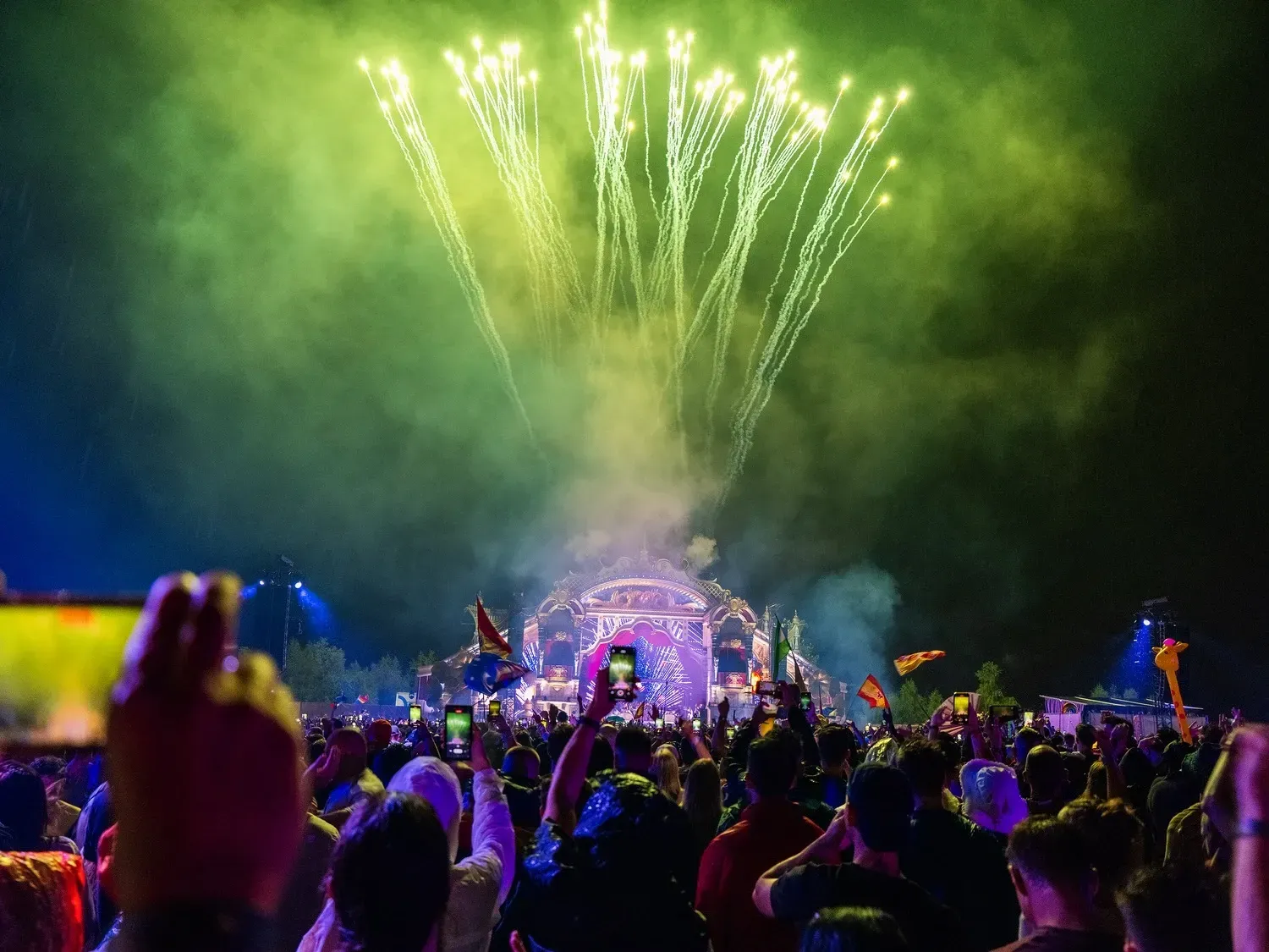 Crowd at outdoor music festival watches fireworks over a stage at night.