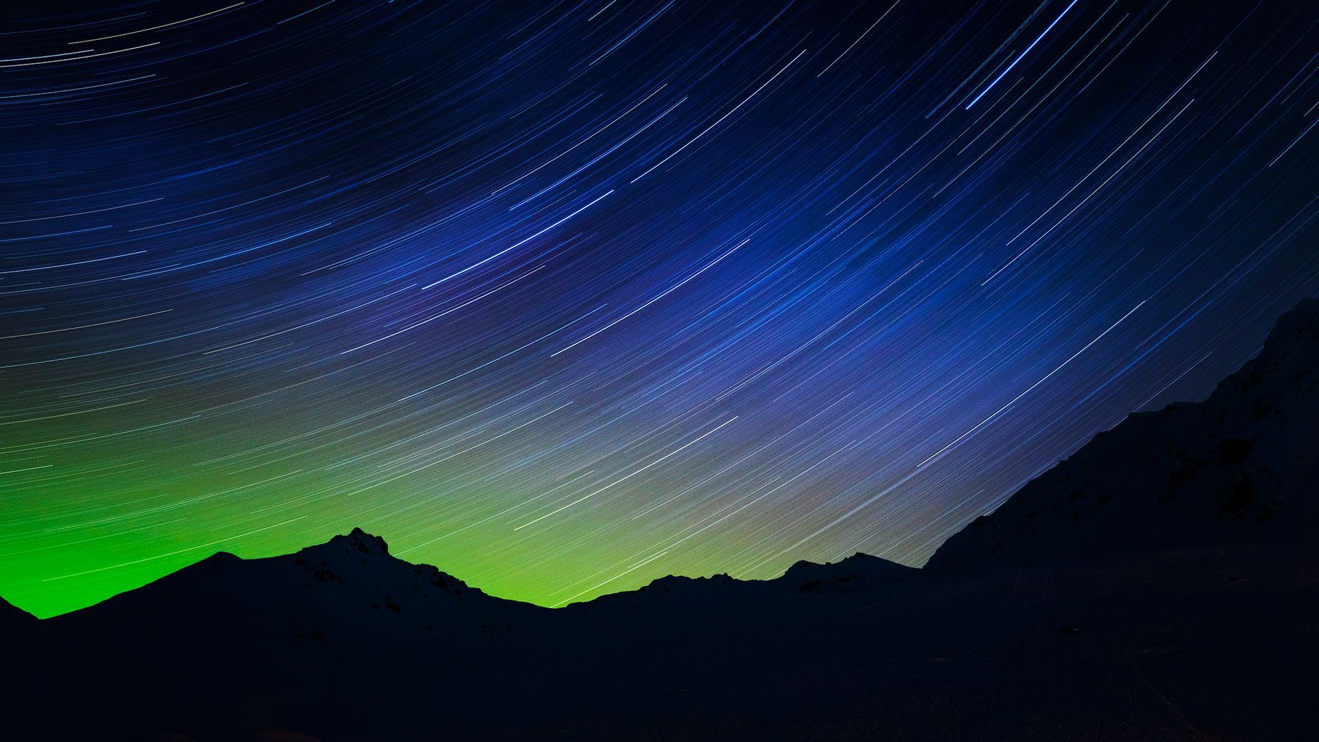 Star trails streak across a blue and green night sky above silhouetted mountains.