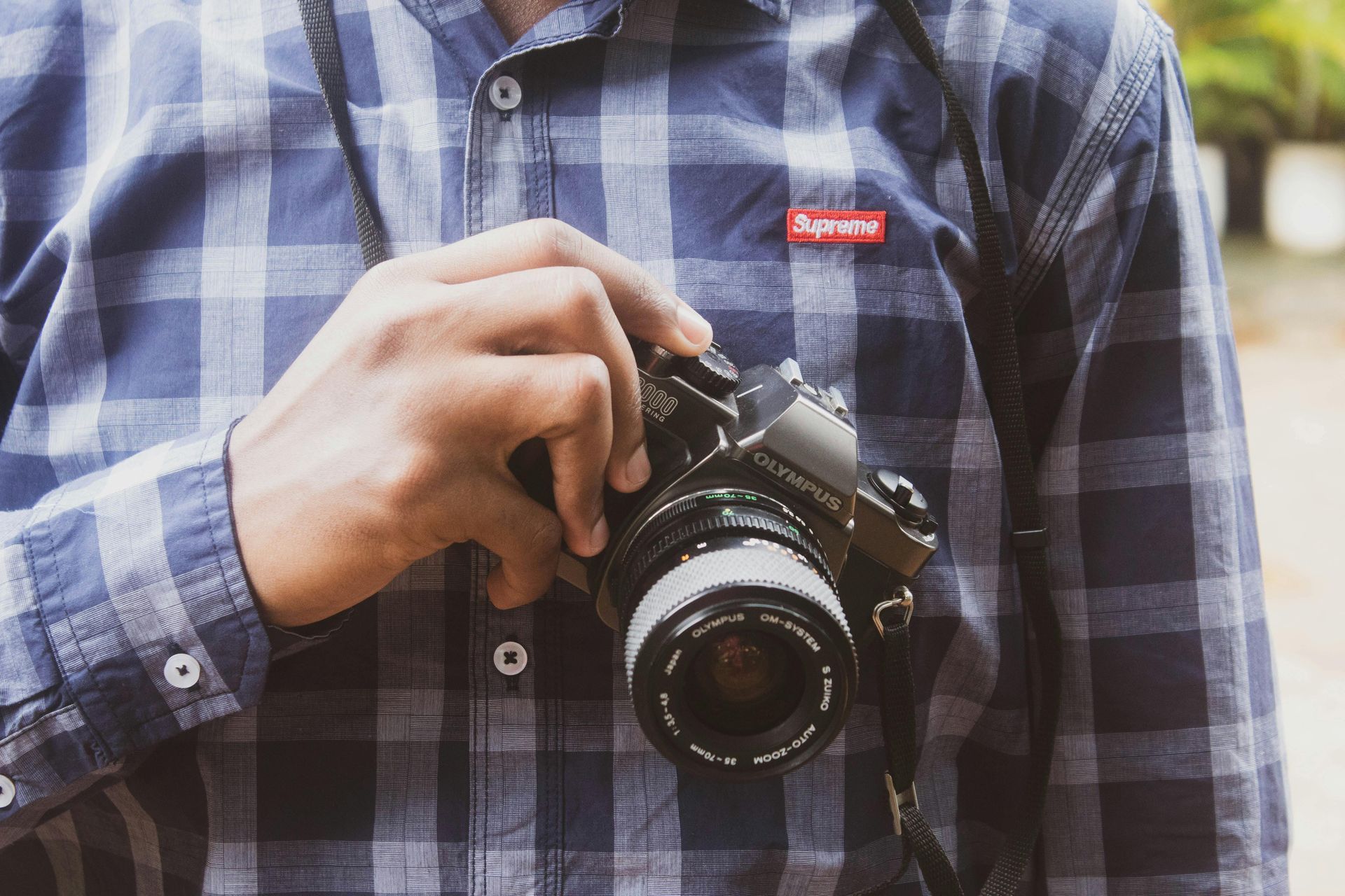 Person holding a black camera, wearing a blue plaid shirt with a red logo.
