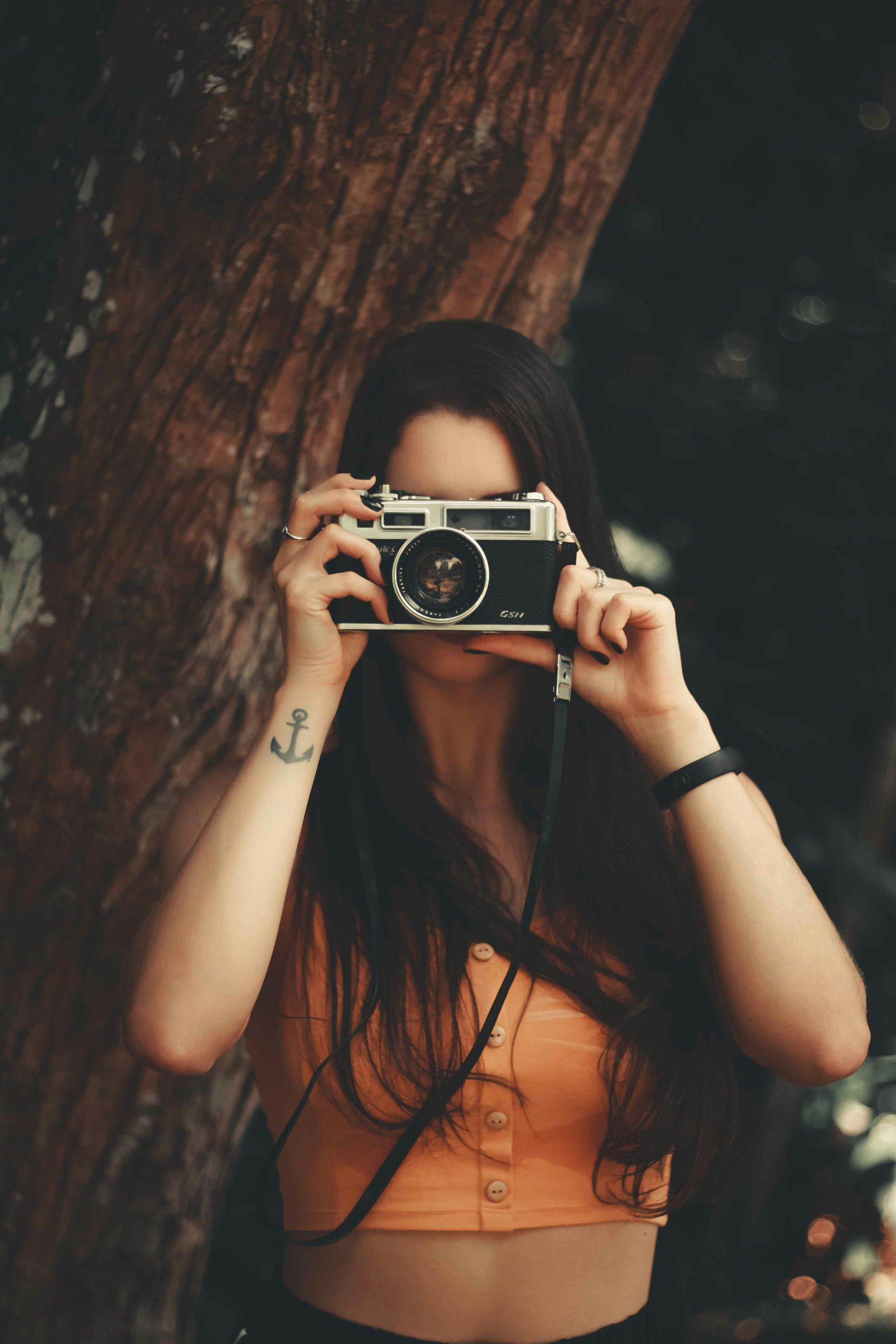 Woman holding a vintage camera, taking a picture outdoors; tree in the background. She wears orange top and has an anchor tattoo.