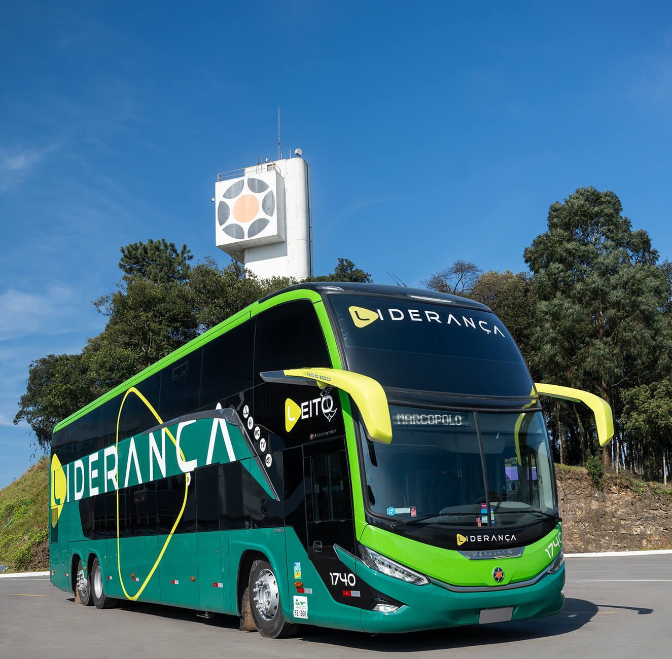 Ônibus Liderança de dois andares, verde e preto, estacionado sob um céu azul, em frente a uma torre branca.