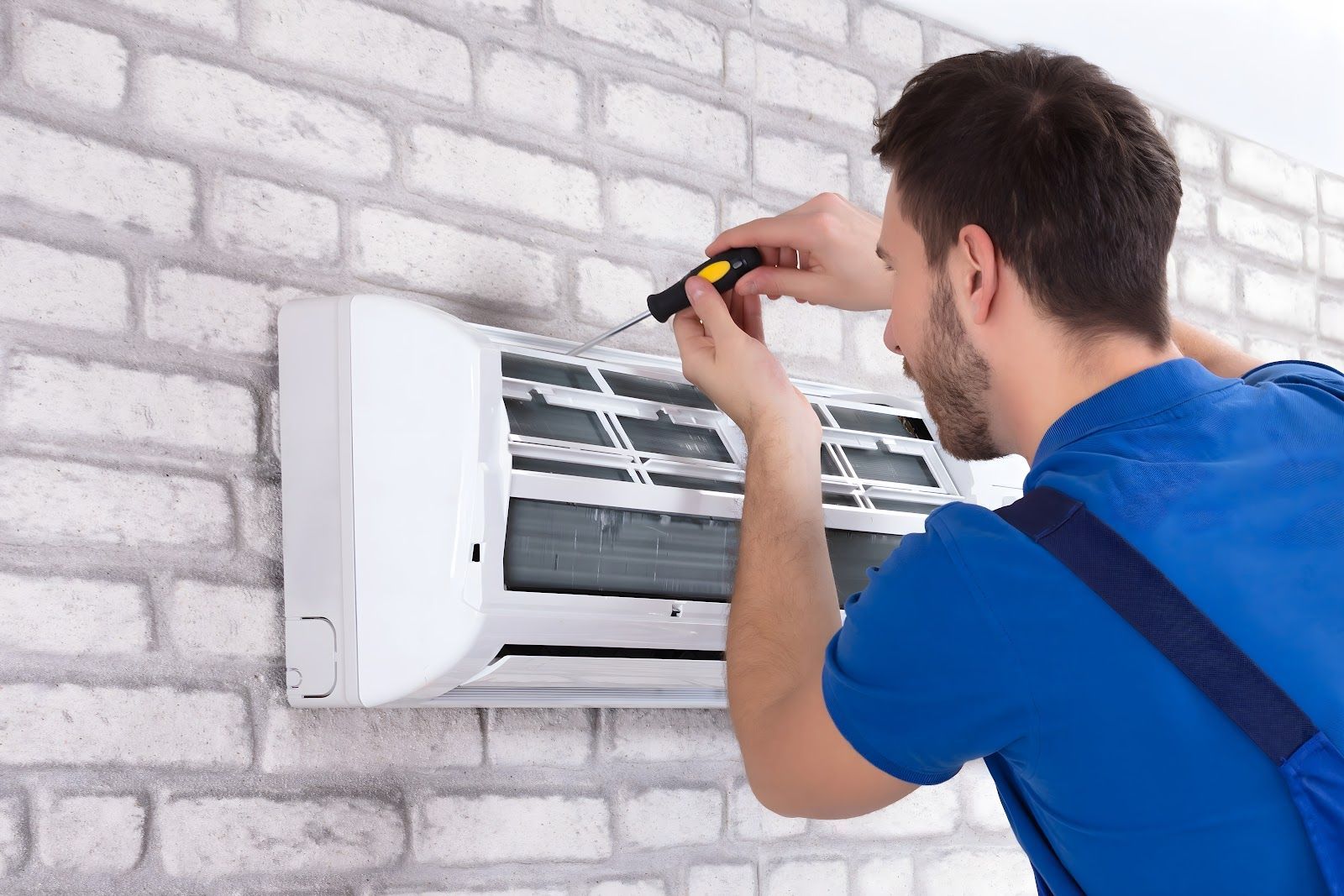 HVAC technician using a screwdriver to service a white wall-mounted air conditioner on a brick wall.