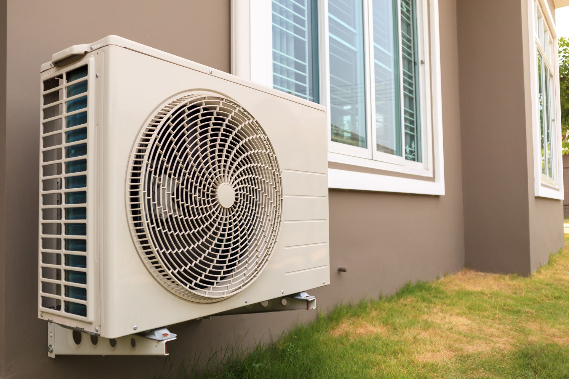 Outdoor air conditioner unit mounted beside a house wall and windows, above a grassy yard.